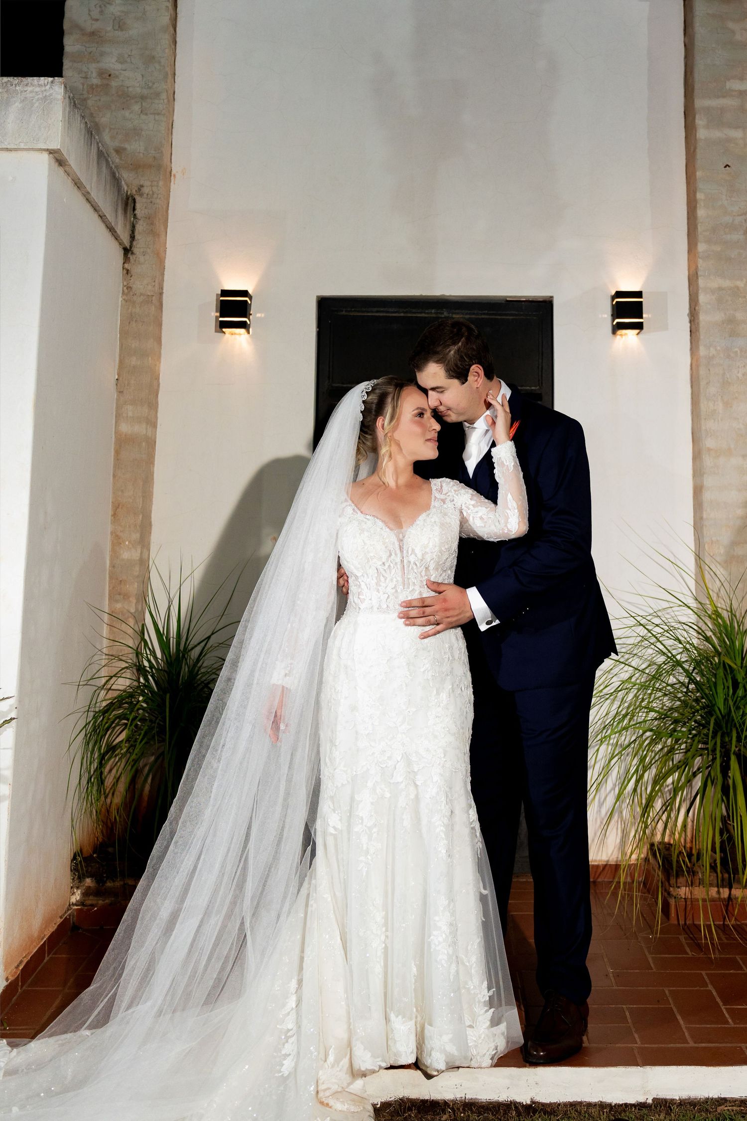 Bride in lace gown with veil and groom in navy suit share romantic moment at night outside elegant venue.