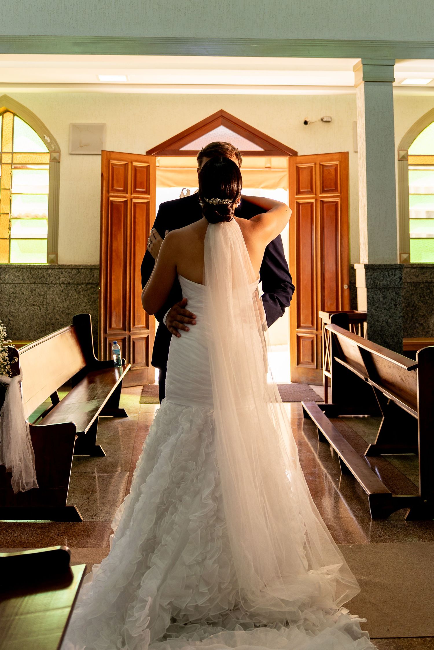 Bride and groom embracing at church altar, bride wearing elegant white mermaid gown with veil, wooden pews lining aisle.