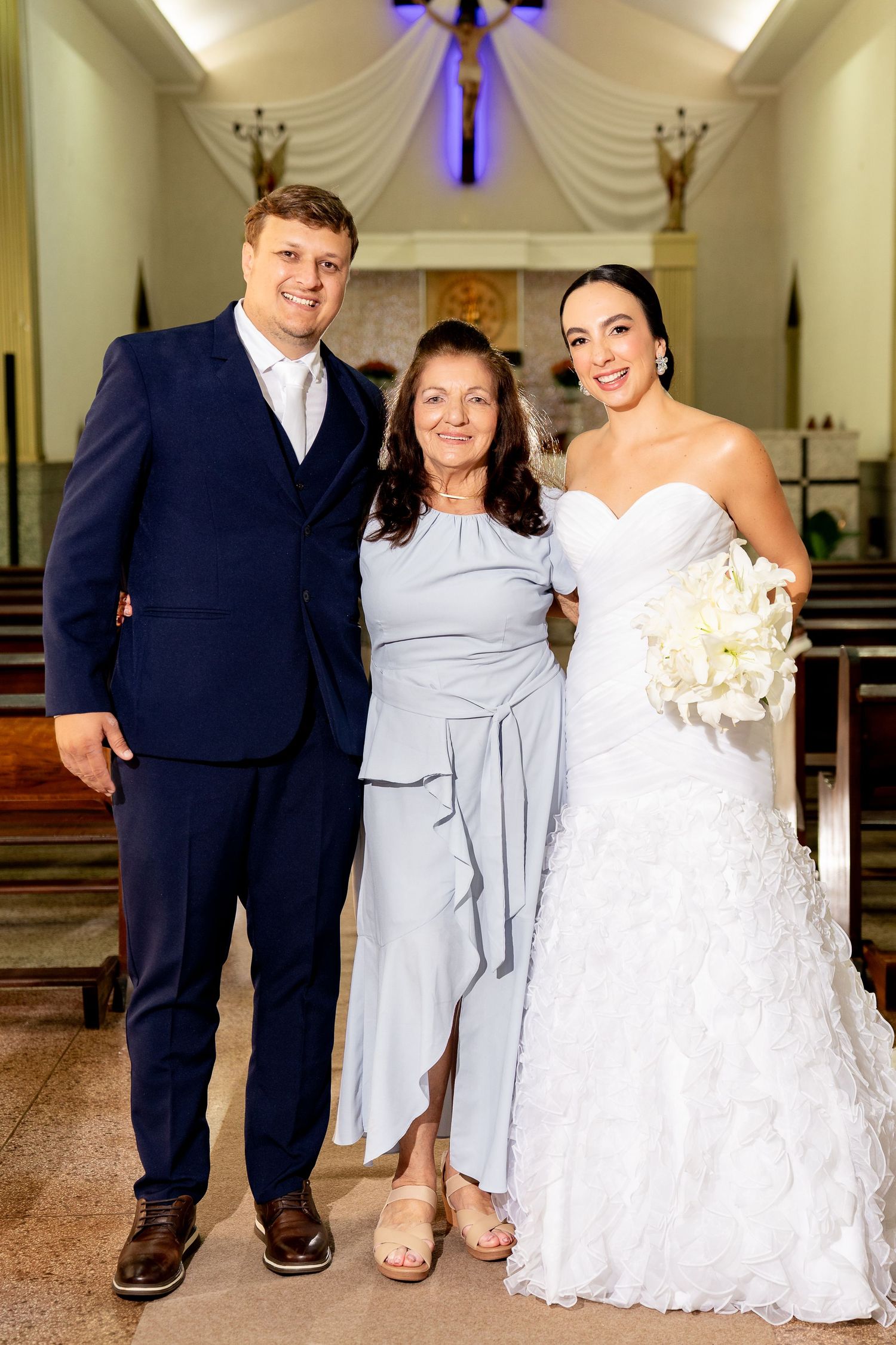 Bride in white gown and groom in navy suit pose with older woman in light blue dress inside a church.