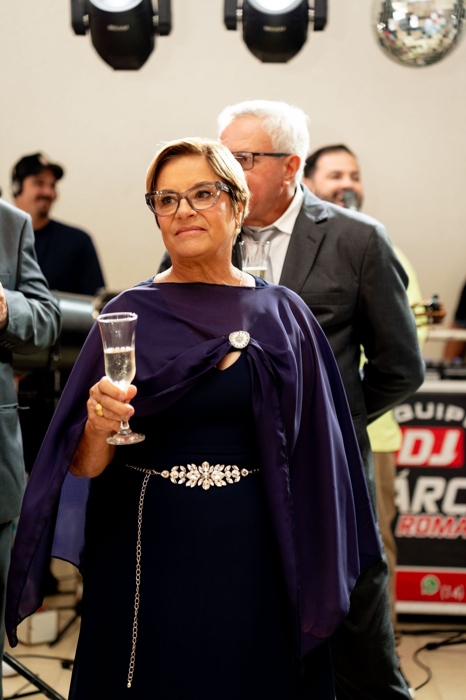 Elegant woman in purple gown holding champagne glass at formal event with DJ setup and guests in background.