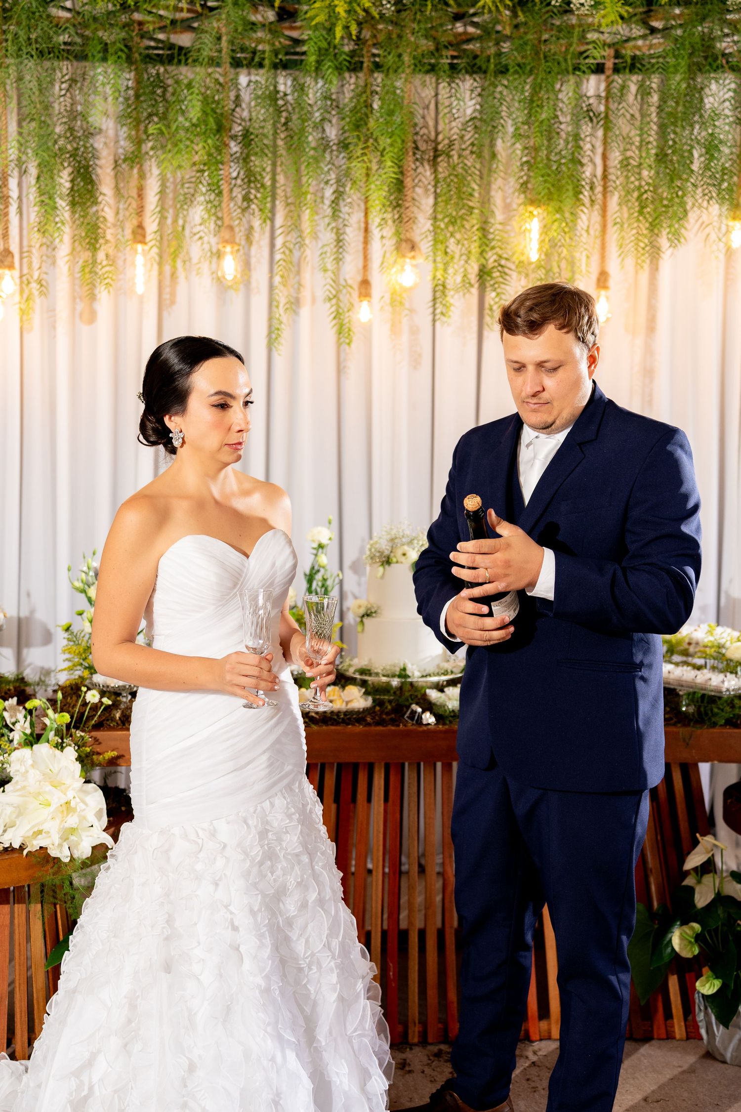 Bride in white gown and groom in navy suit opening champagne bottle at elegant wedding reception with greenery decor.