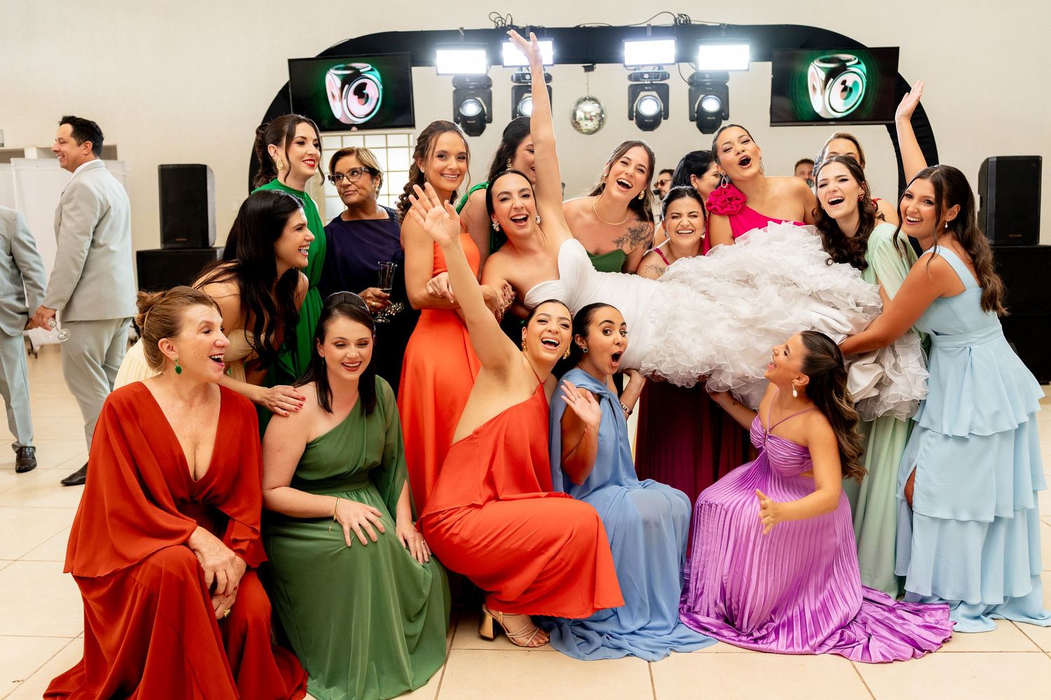 Bride tossed in air by bridesmaids in colorful gowns celebrating at wedding reception party.