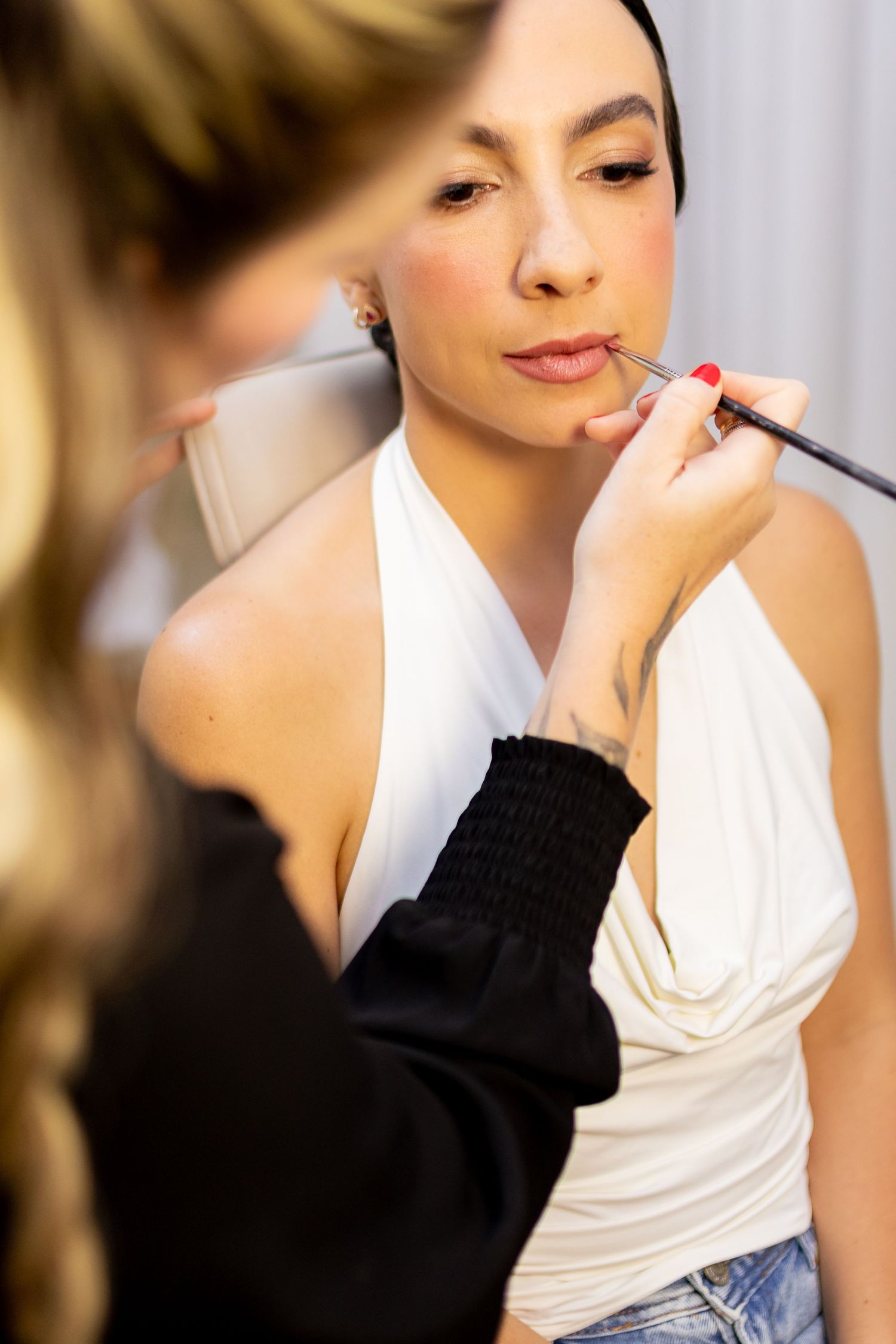 Makeup artist applying lip color to a woman in a white halter top using a professional brush.