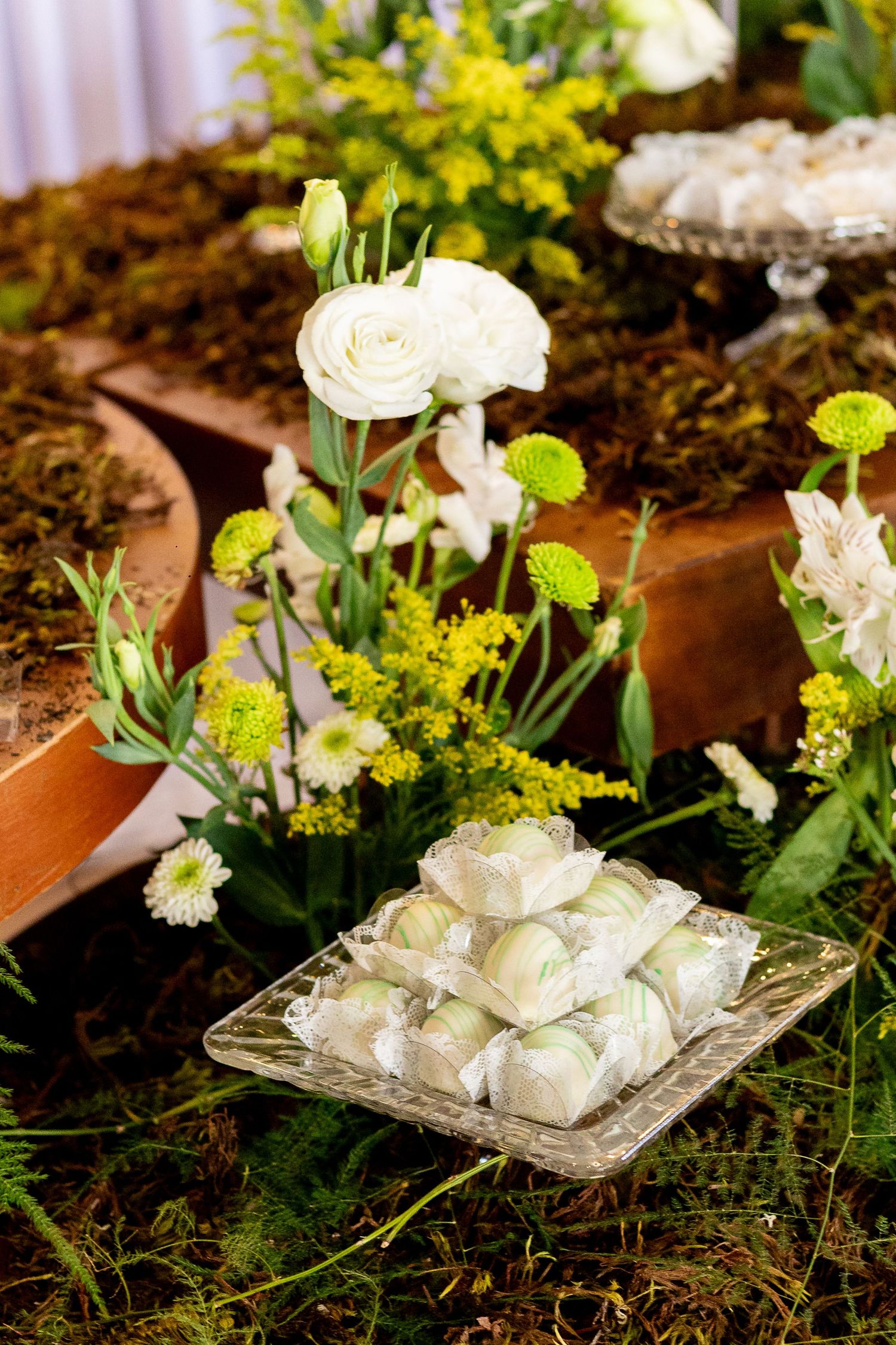 Elegant wedding dessert table with white roses, yellow flowers, and white candy treats on a glass plate amid green moss.