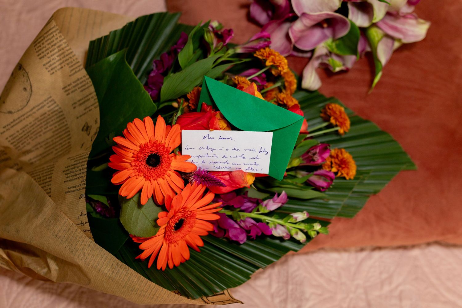 Colorful bouquet of orange gerberas, purple flowers, and greenery wrapped in brown paper with a handwritten note card.