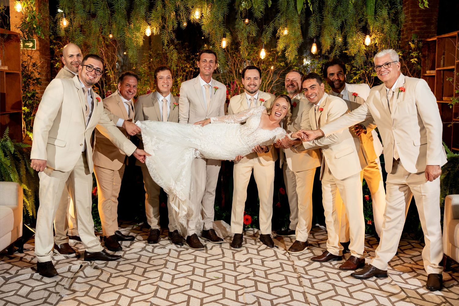 Groom and bride lifted by groomsmen in tan suits at elegant outdoor wedding reception with string lights.