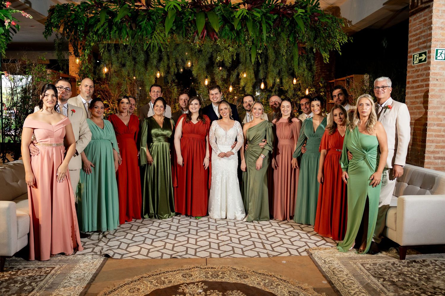 Wedding party group photo with bridesmaids in red and green gowns flanking bride in white dress indoors.