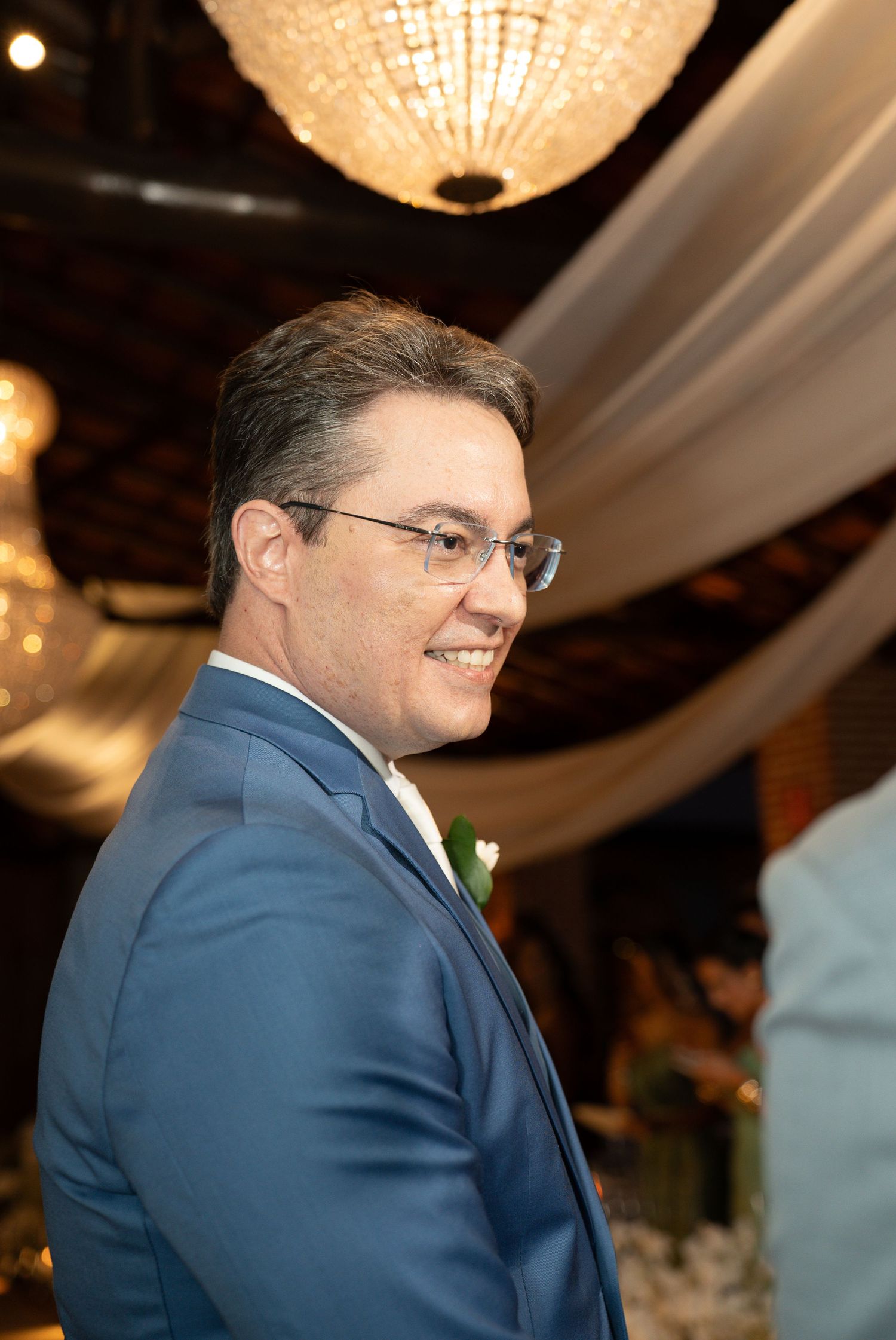 Smiling man in blue suit and glasses at elegant wedding reception with chandelier lighting.