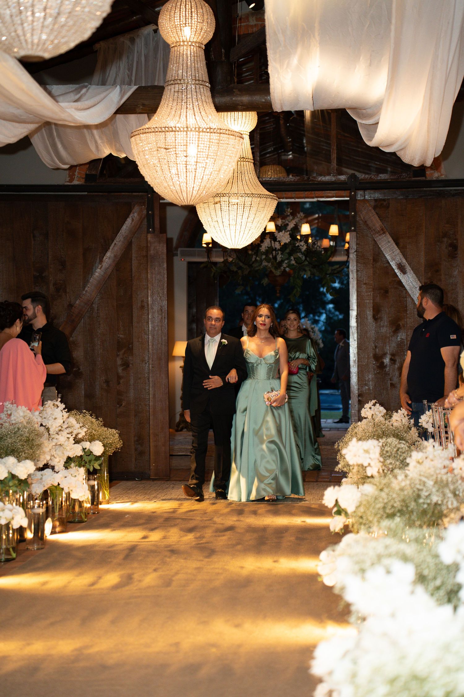 Elegant wedding ceremony in a rustic barn with chandeliers, floral aisle, and guests in formal attire walking the aisle.
