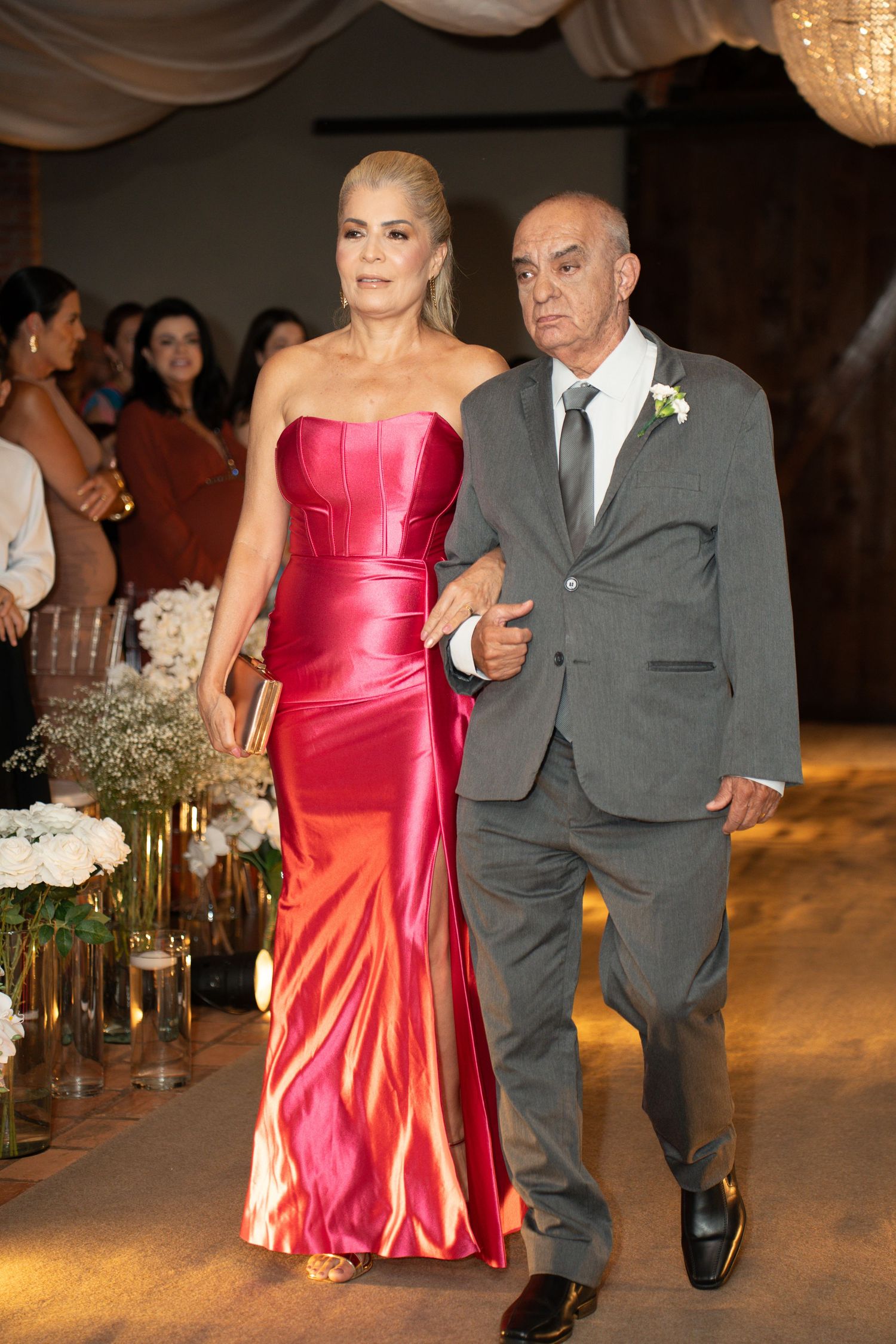 Elegant couple walking down wedding aisle, woman in red satin gown, man in gray suit with boutonniere.