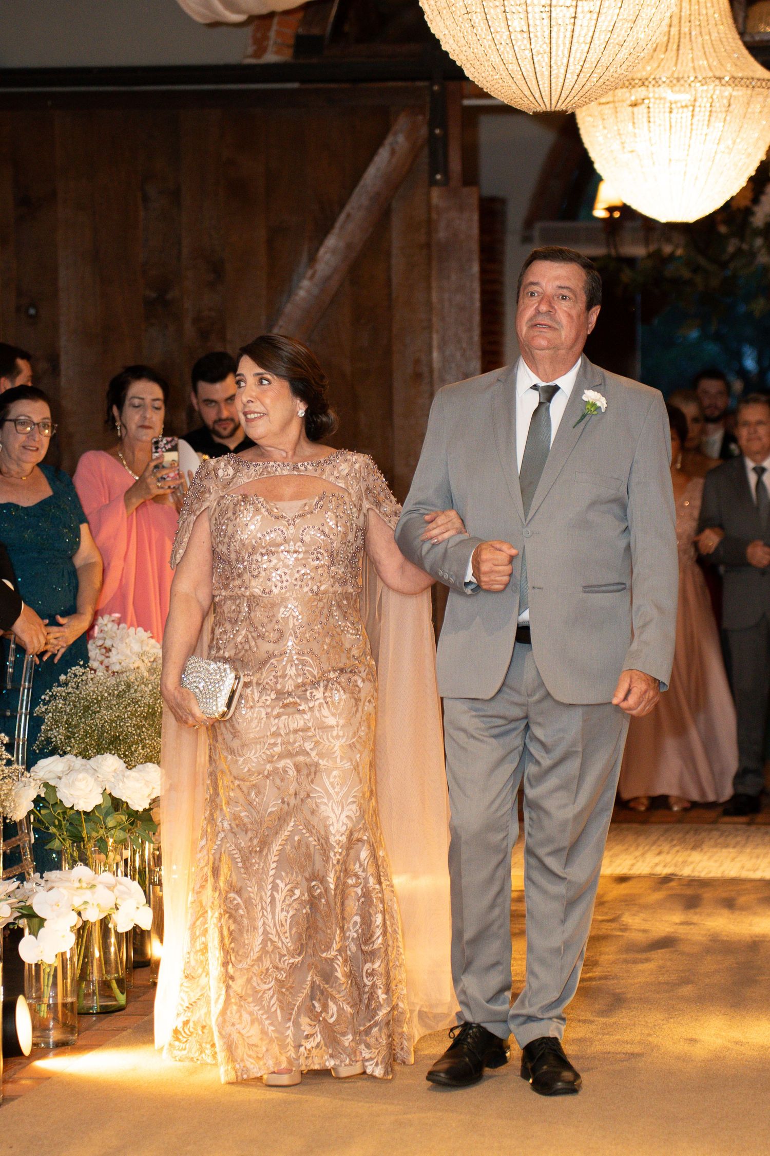 Elegant couple walking down wedding aisle, woman in gold sequin gown, man in gray suit with boutonniere, guests watching.
