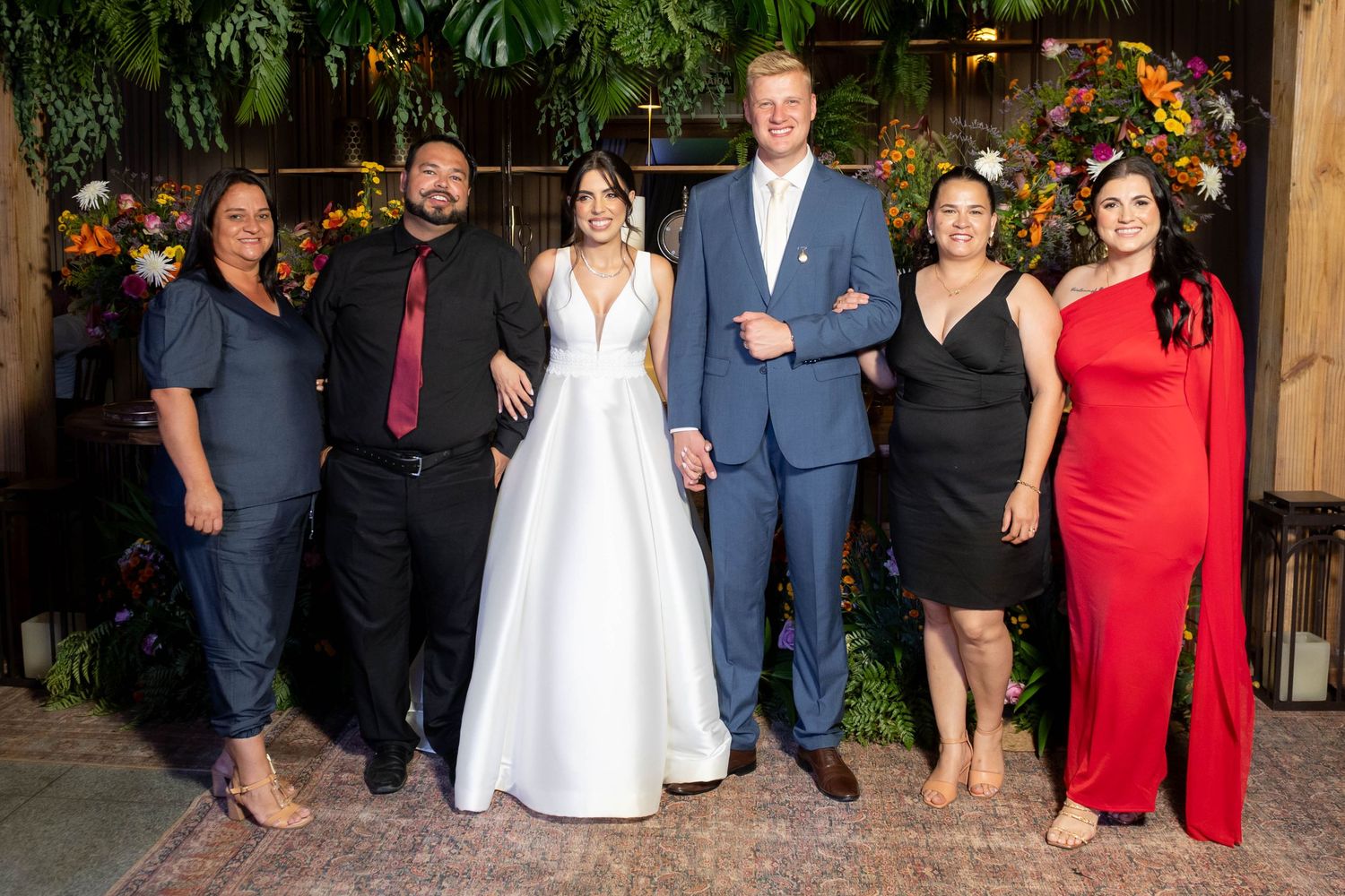Wedding party of six posing together, bride in white gown and groom in suit, flanked by guests in formal attire.