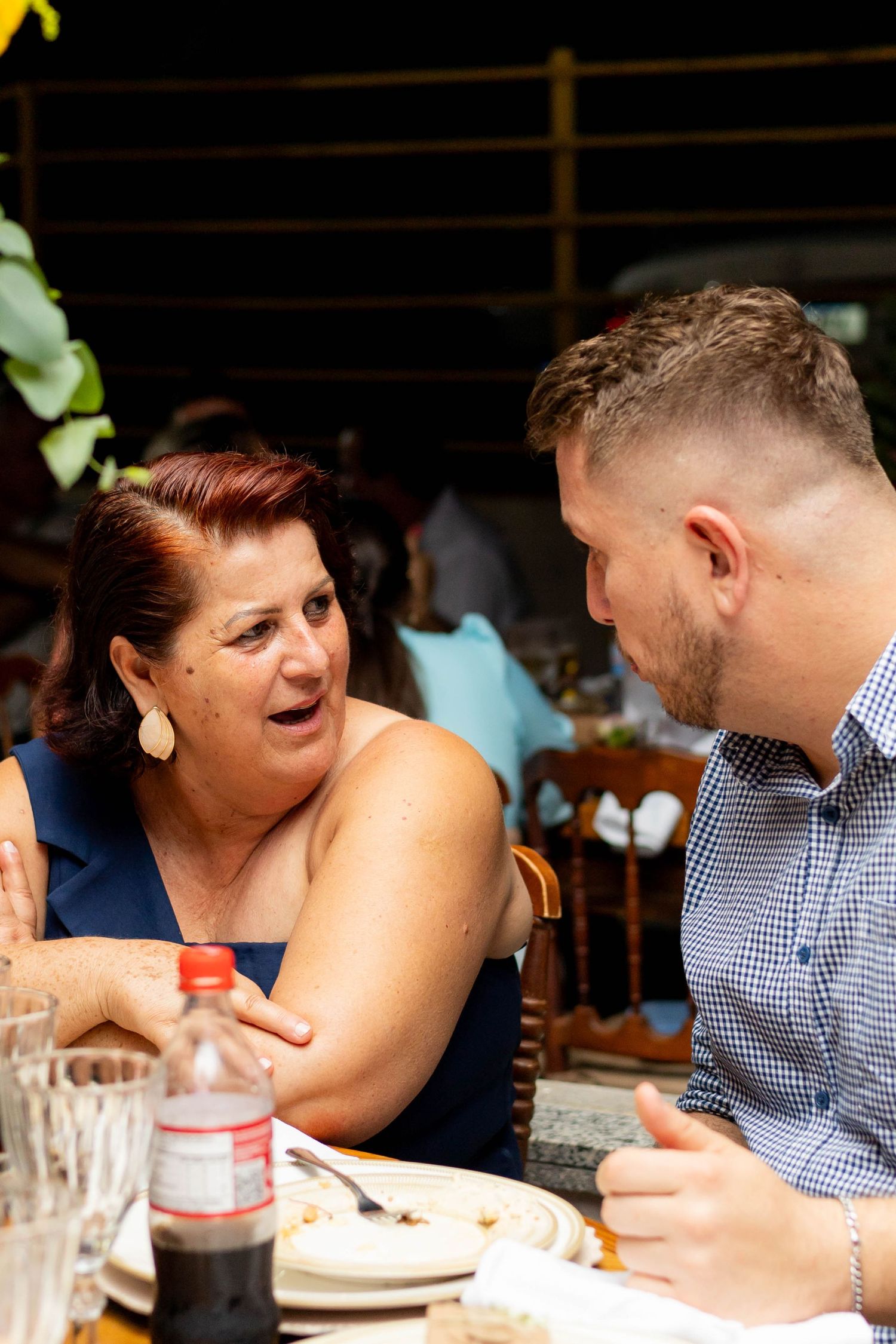 Older woman with red hair in blue dress talking to young man in checkered shirt at outdoor restaurant table.