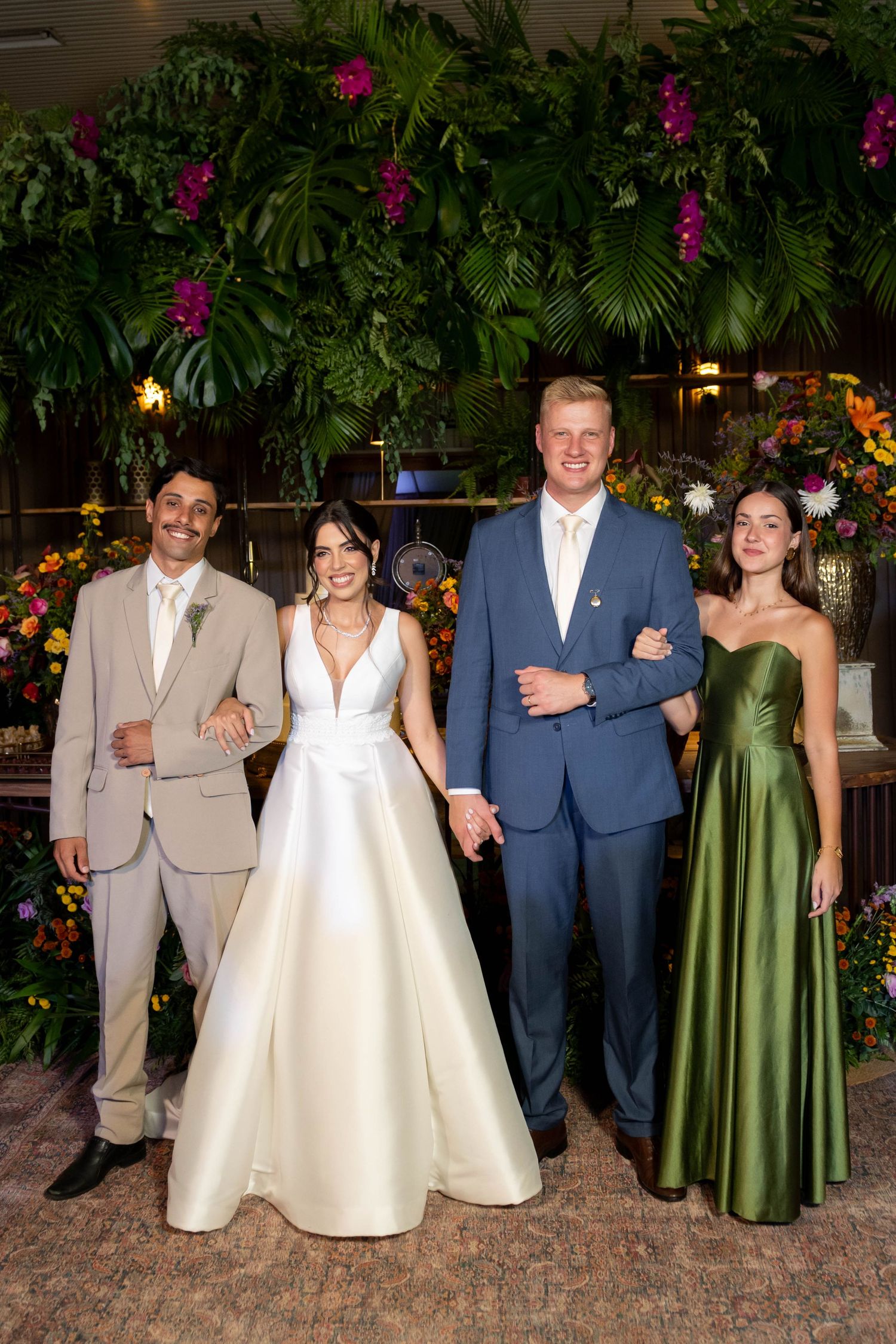 Wedding party of four posing in front of lush tropical greenery, bride in white gown, groom in blue suit.