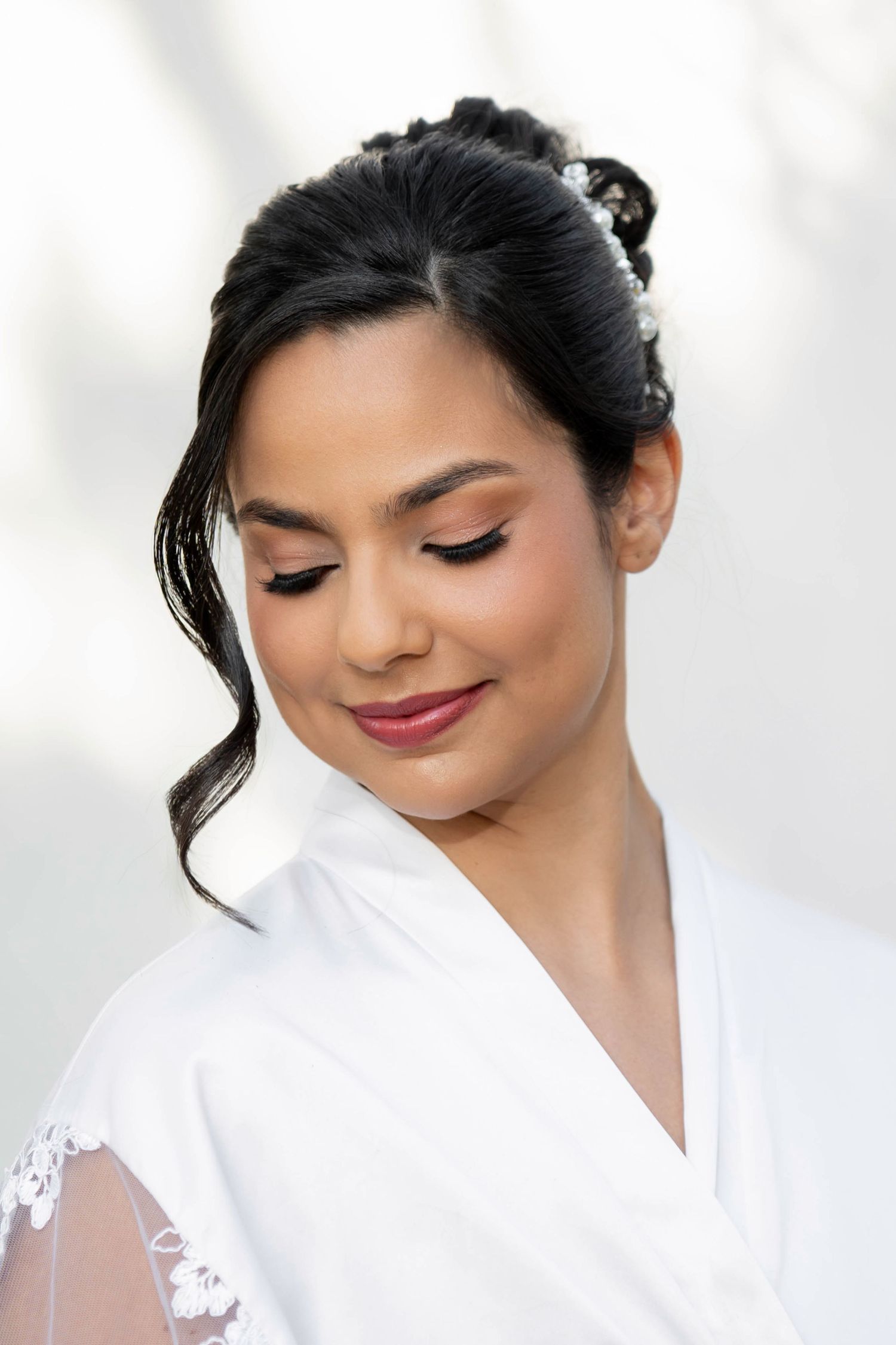 Bride with elegant updo hairstyle, floral hair accessories, and natural makeup wearing a white lace robe.