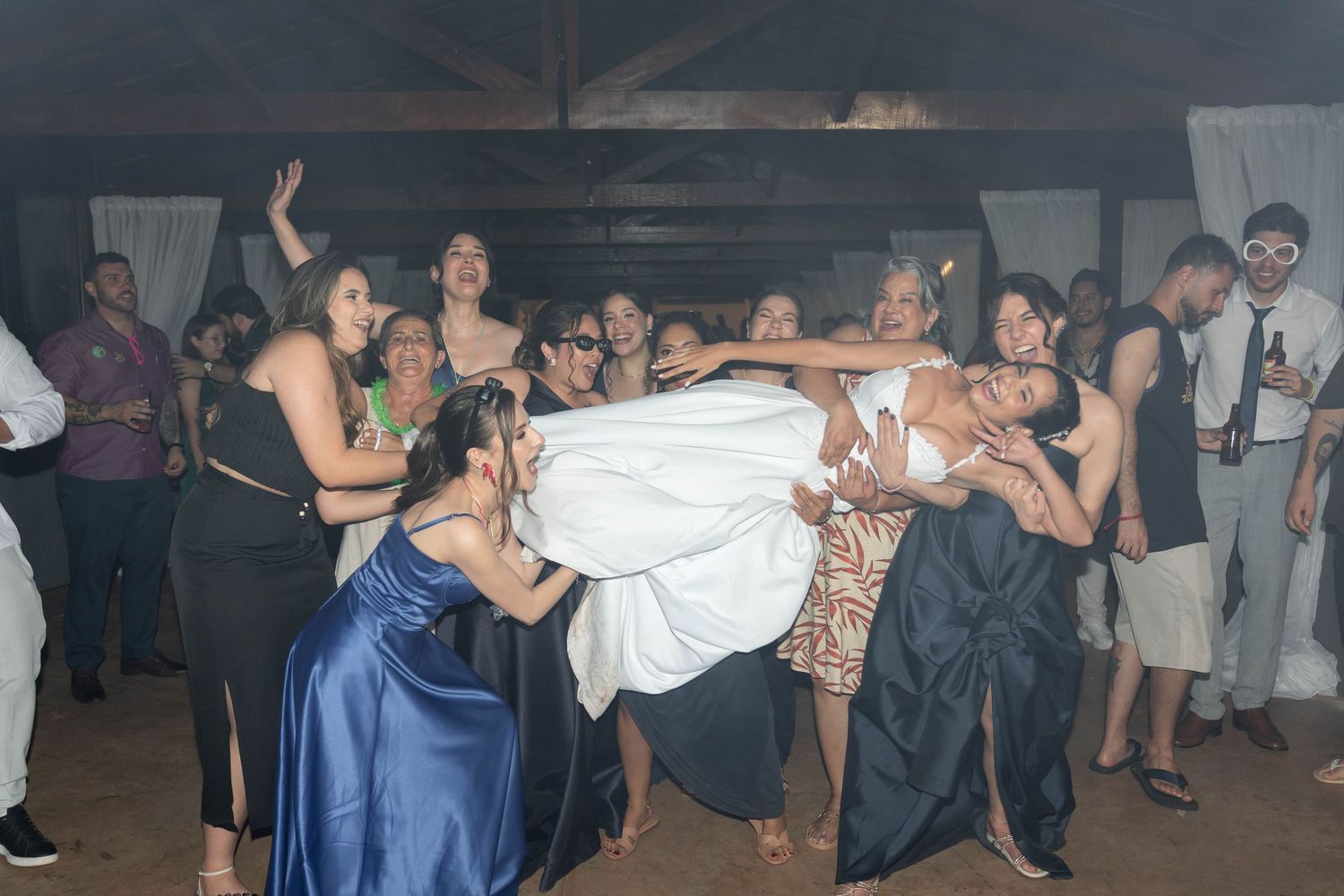 Bride being lifted by wedding guests on dance floor during lively indoor wedding reception celebration.