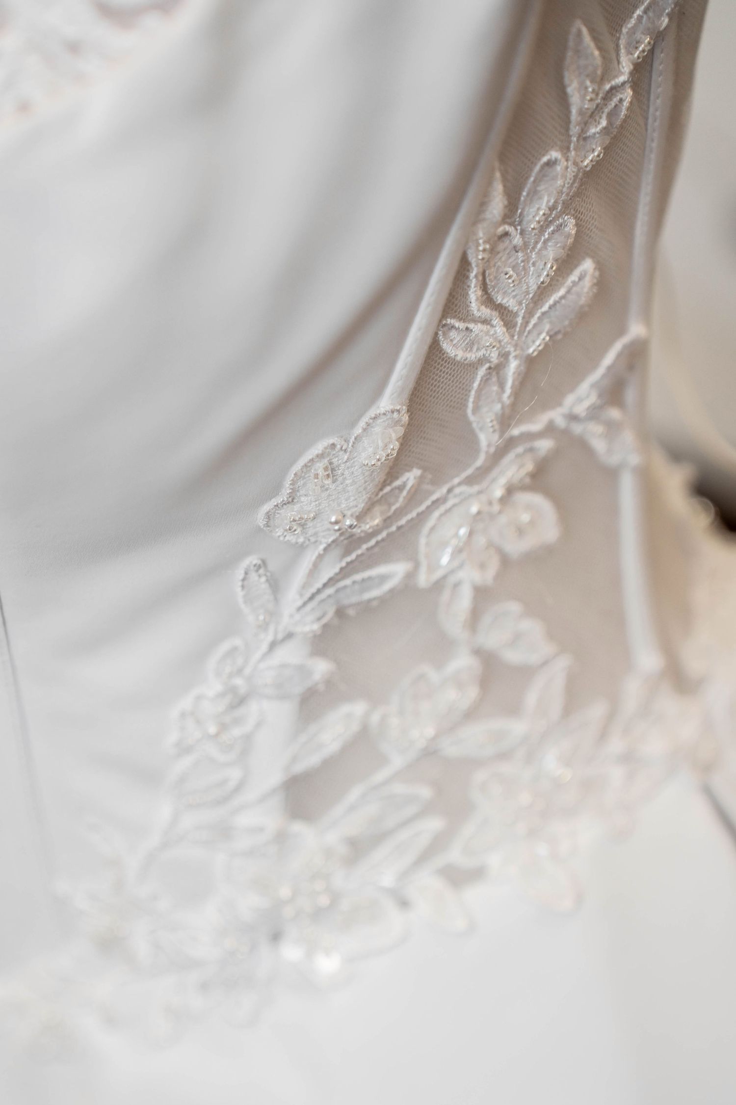 Close-up of elegant white wedding dress lace detail featuring delicate floral and leaf embroidery with beading.