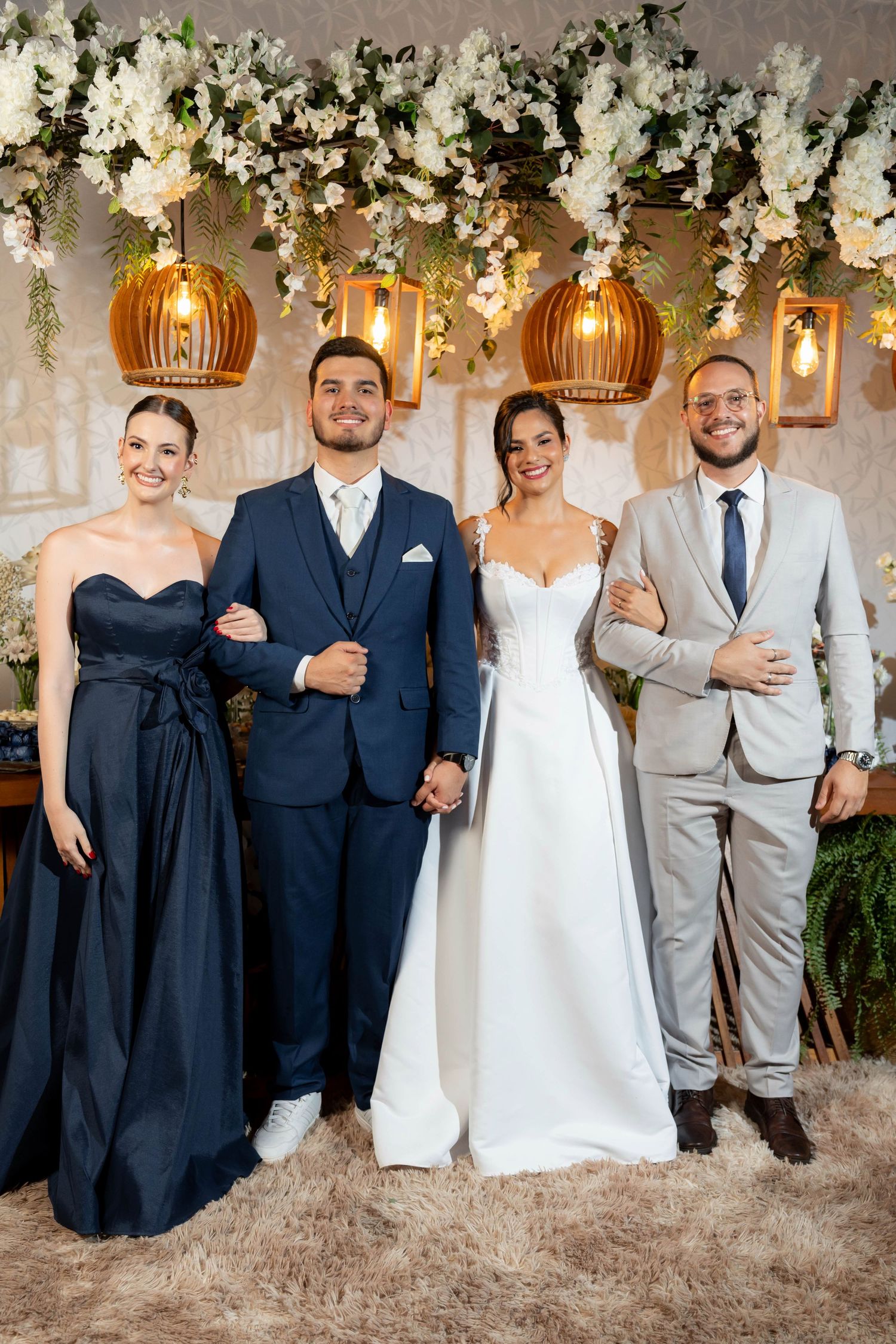 Wedding party of four posing elegantly under floral decor and warm pendant lights, bride in white gown and groom in navy suit.