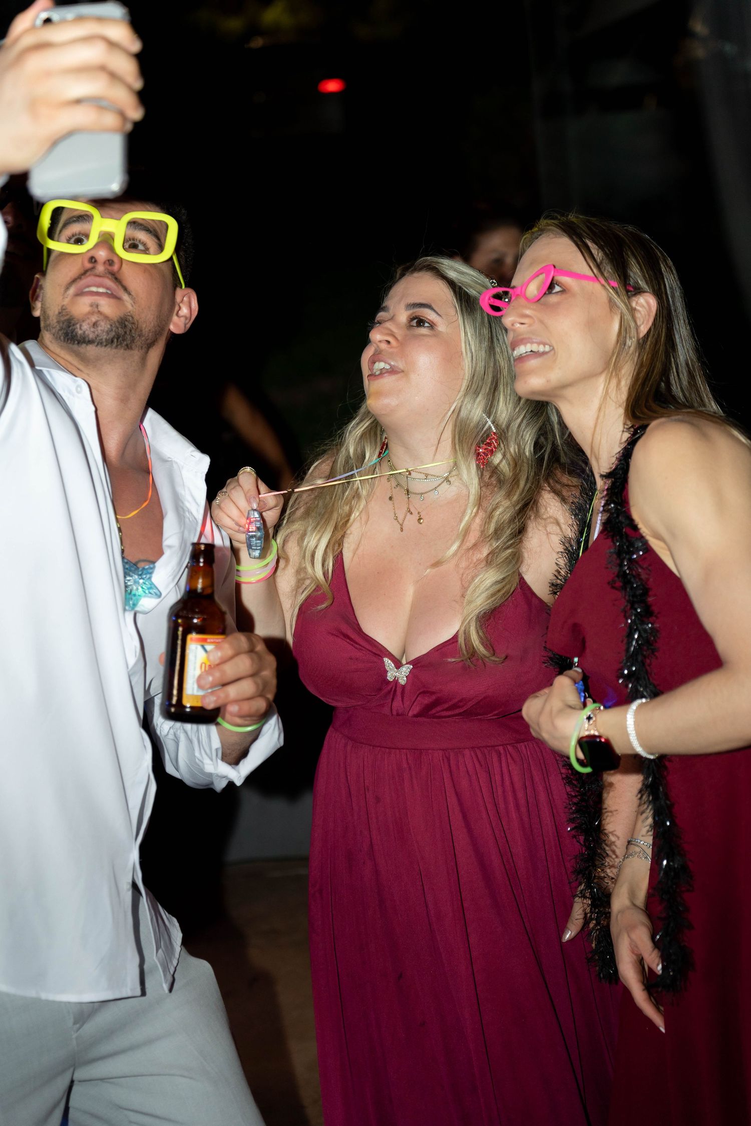 Three party guests in formal wear take a selfie, wearing colorful novelty glasses at a festive evening event.