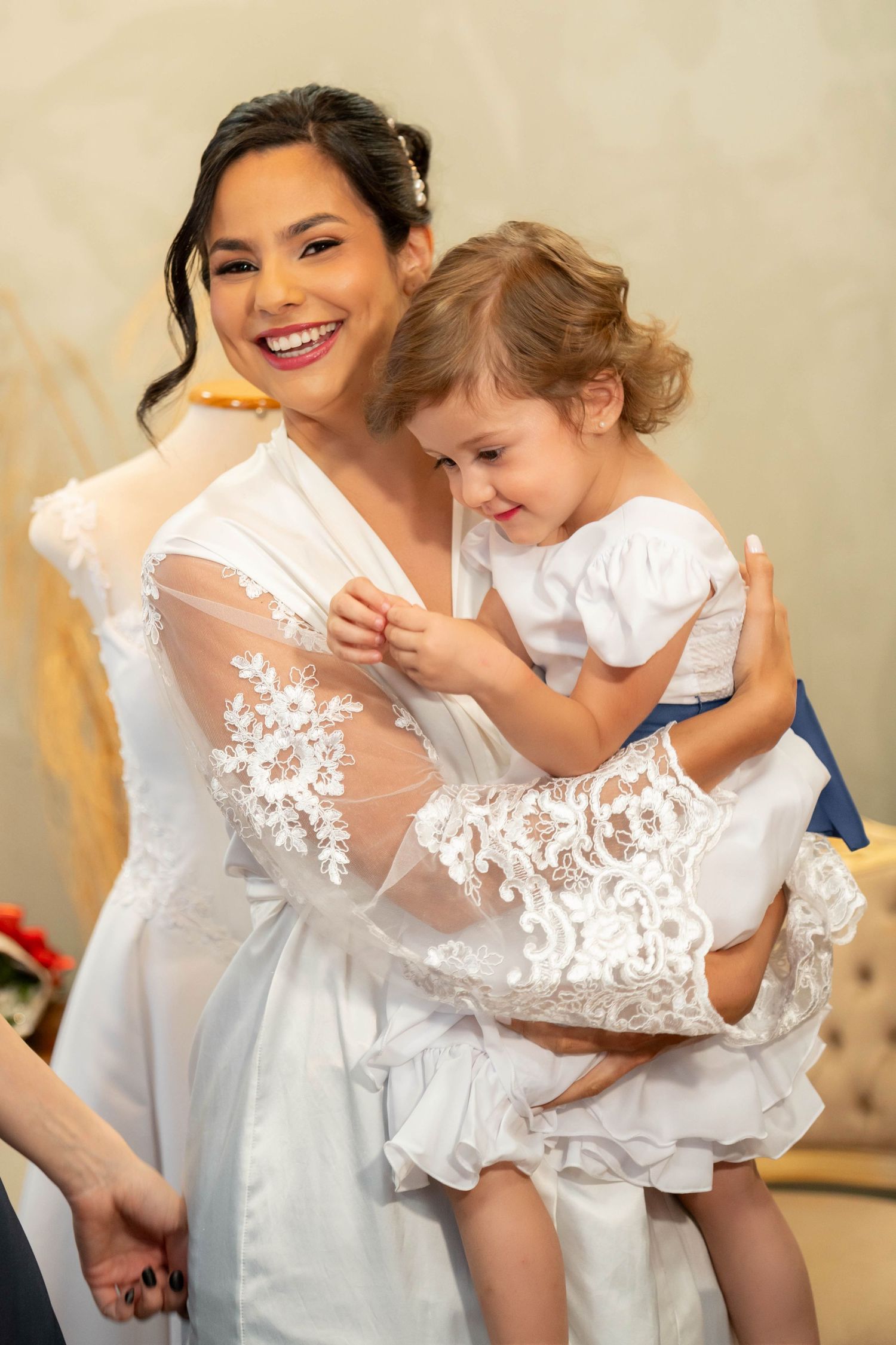 Smiling bride in white lace robe holds a young girl in a white dress during wedding preparations.