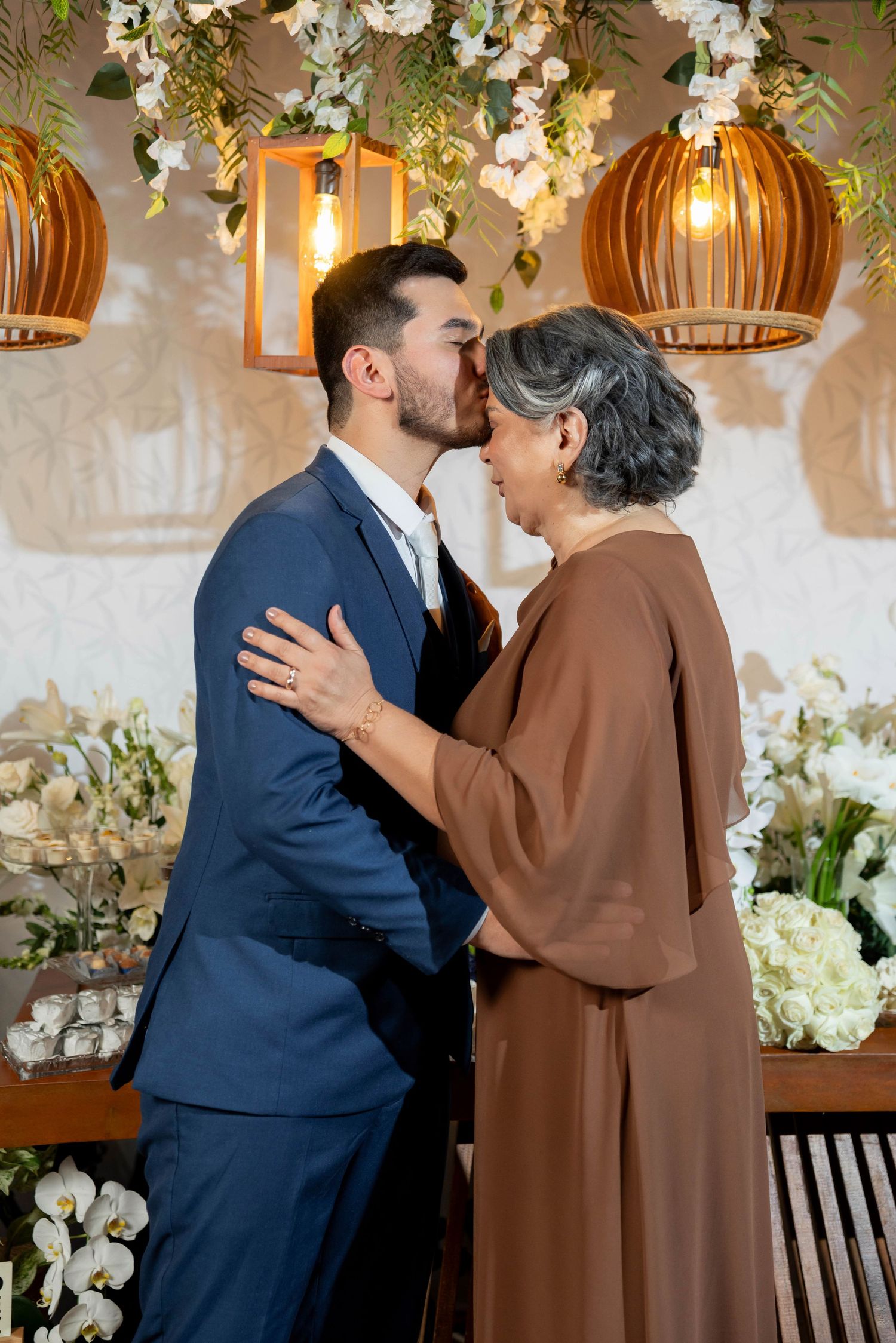 Groom in navy suit shares a kiss with older woman in brown dress at elegant floral wedding reception.