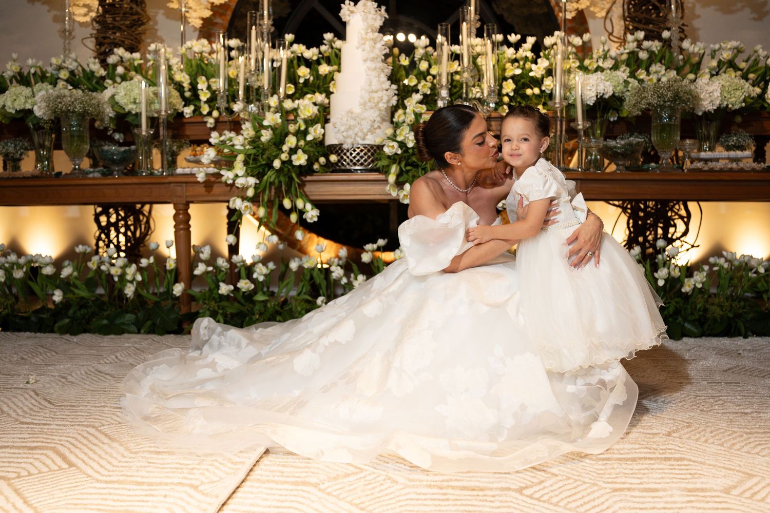 Bride in a white ball gown holding a toddler in white dress at an elegant wedding reception with floral decor.