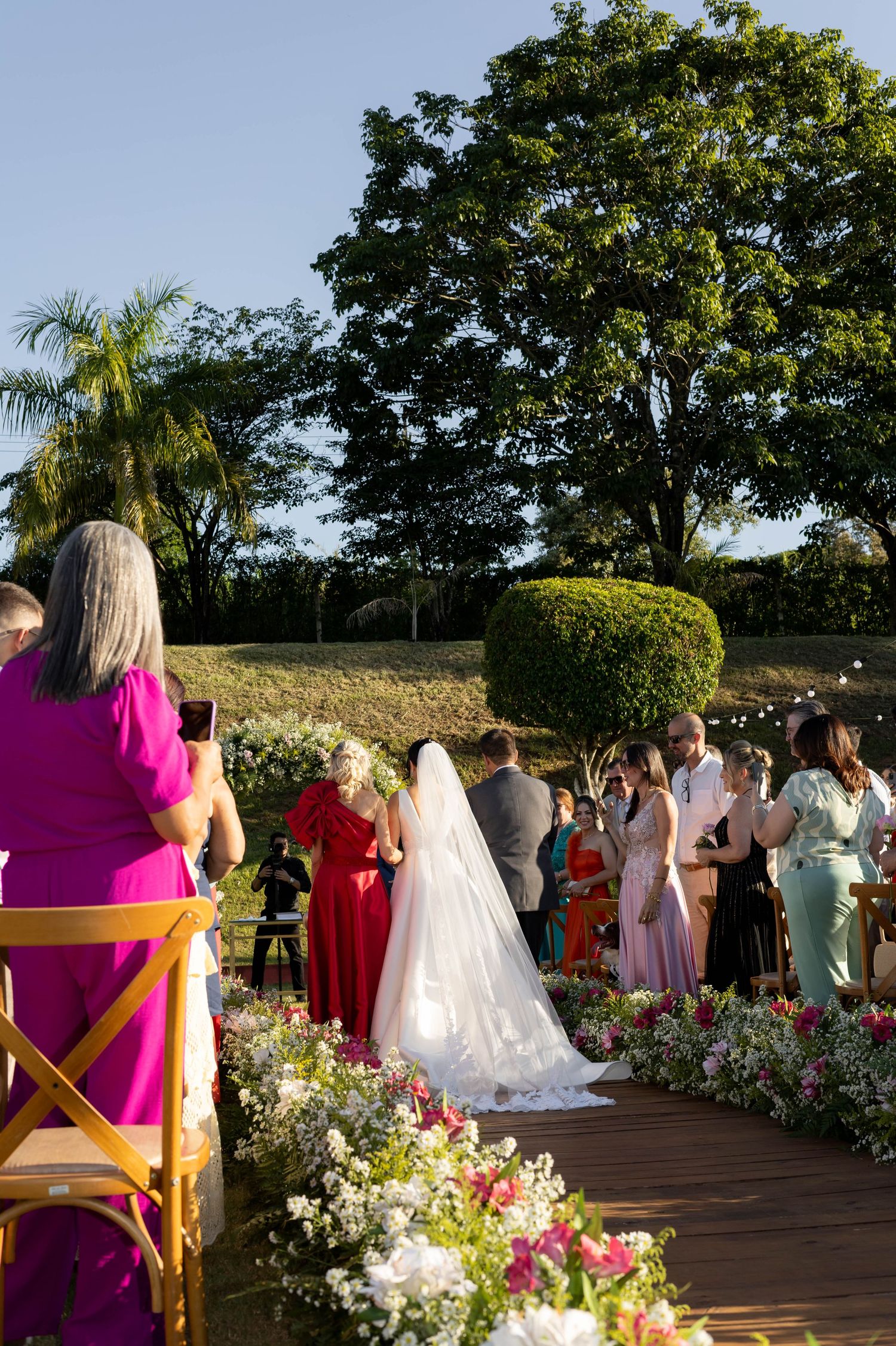 Outdoor wedding ceremony on wooden deck with floral aisle, bride in white veil, guests in colorful attire, lush green trees backdrop.