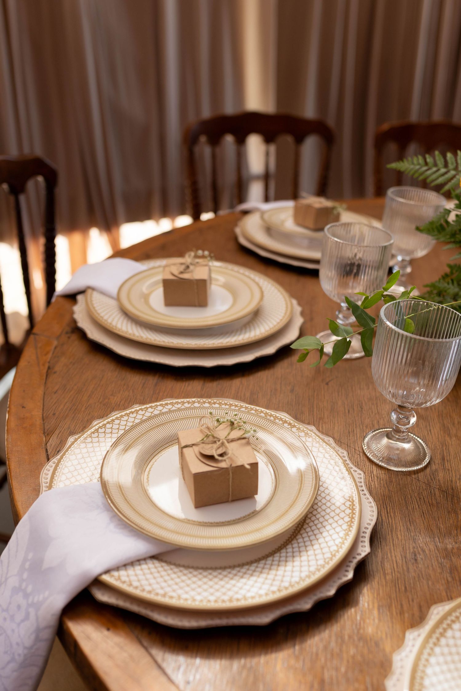Elegant wooden dining table set with gold-rimmed plates, crystal glasses, kraft gift boxes, and green botanical accents.