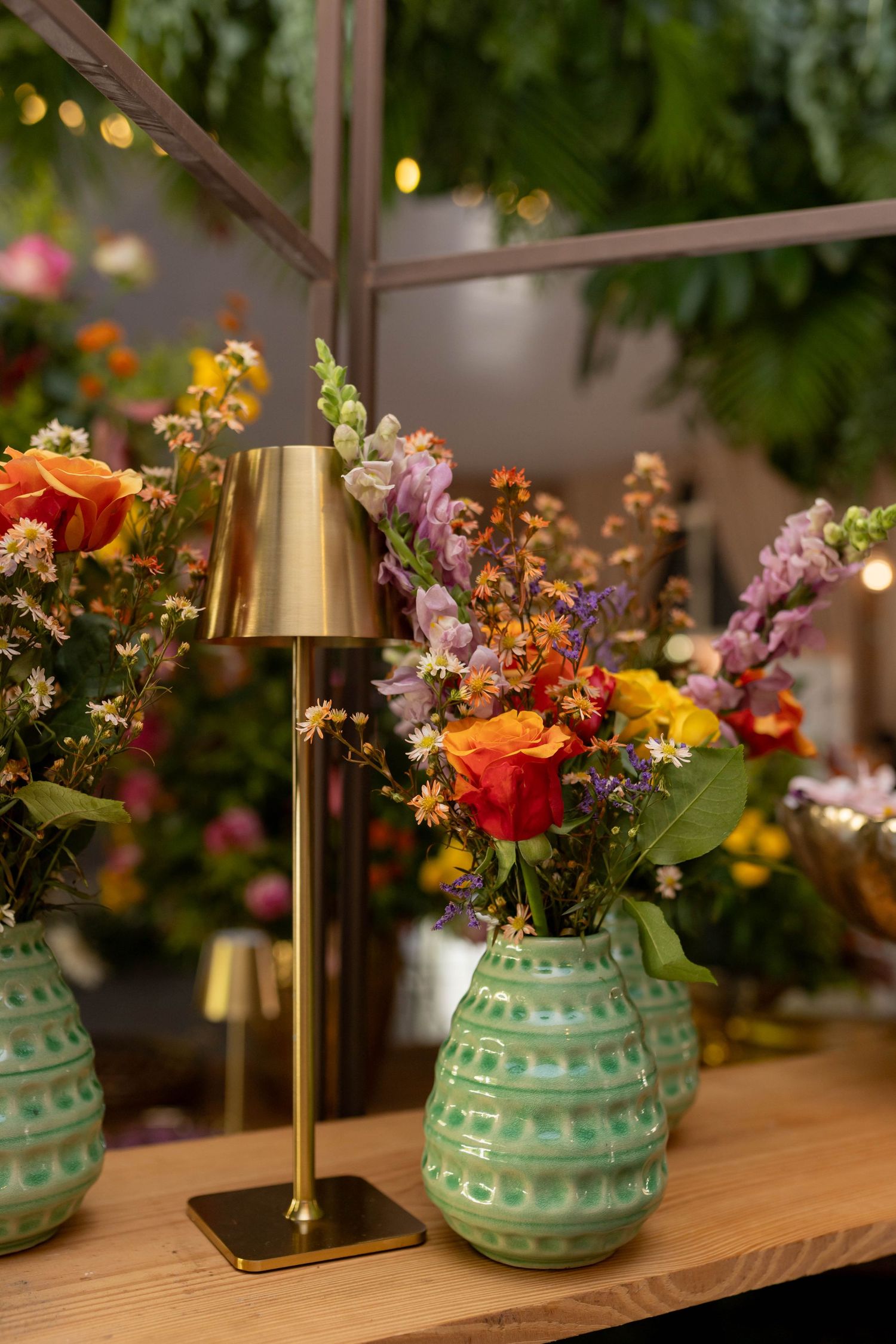 Green ceramic vases with colorful wildflowers beside a gold lamp on a wooden table in a lush floral shop.