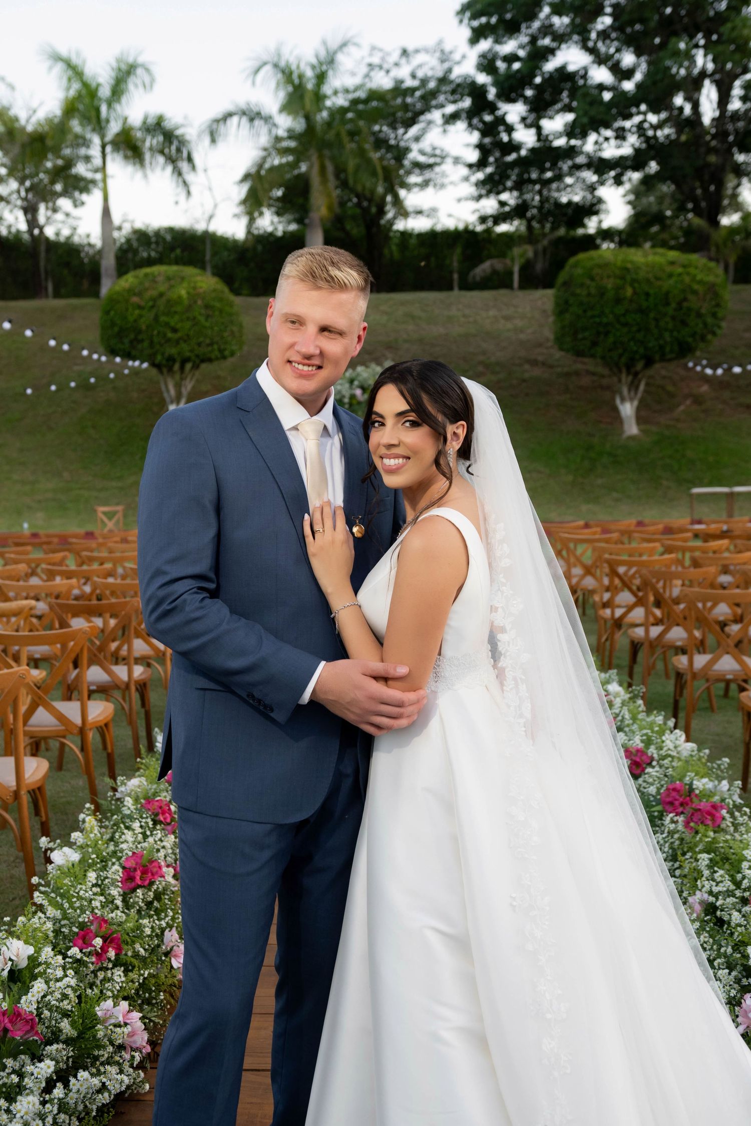 Bride and groom posing at outdoor wedding ceremony with floral arrangements and wooden chairs in lush garden setting.