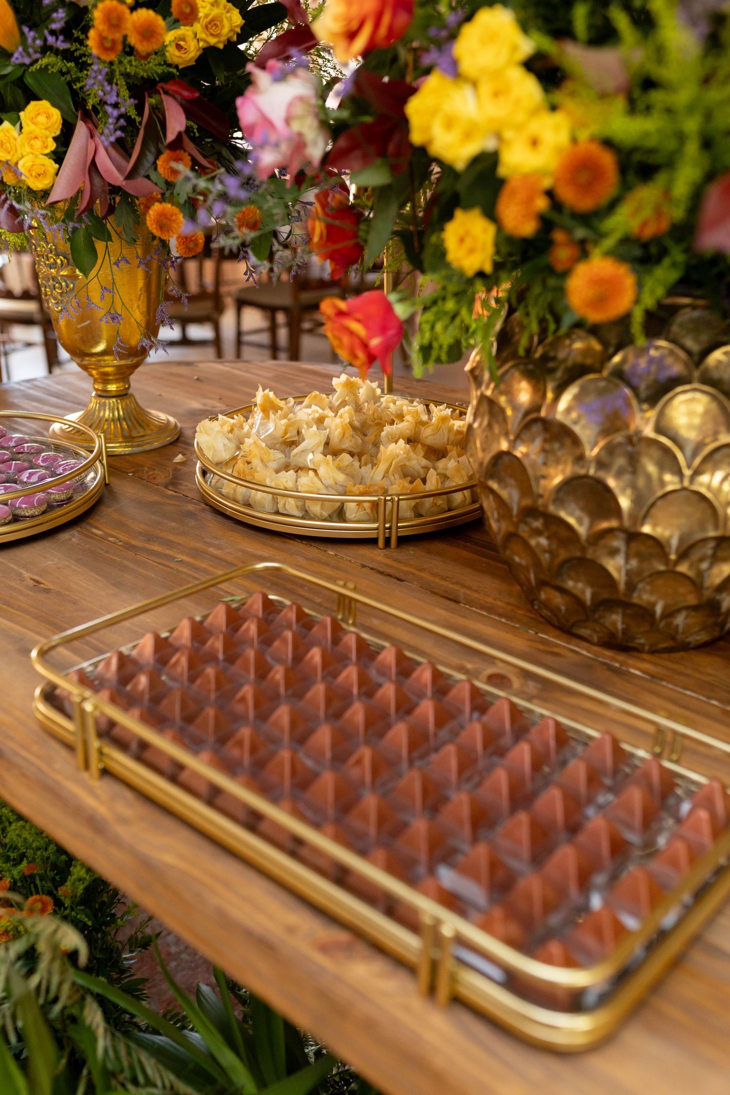 Elegant dessert table with gold trays of chocolates, popcorn, colorful floral arrangements in gold vases on wooden surface.