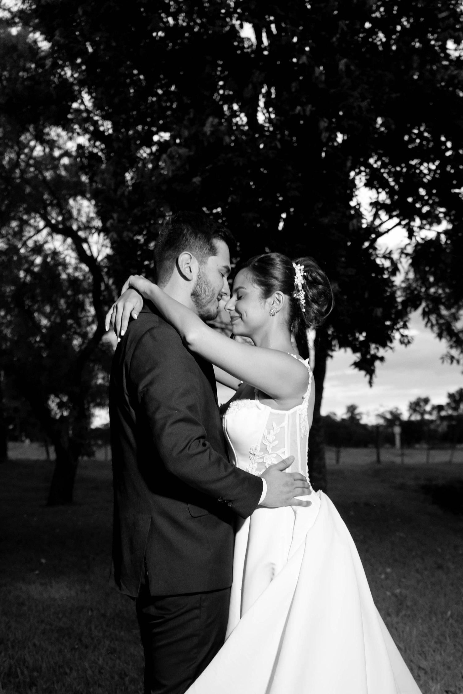 Black and white photo of a bride and groom embracing outdoors near trees at dusk.