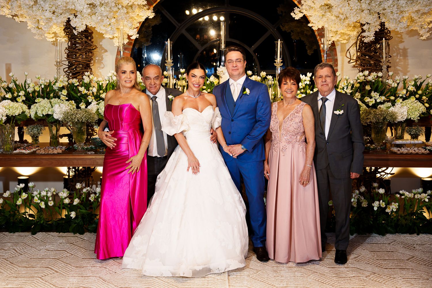 Elegant wedding party poses together in formal attire, featuring bride in white gown and groom in blue suit, surrounded by family.