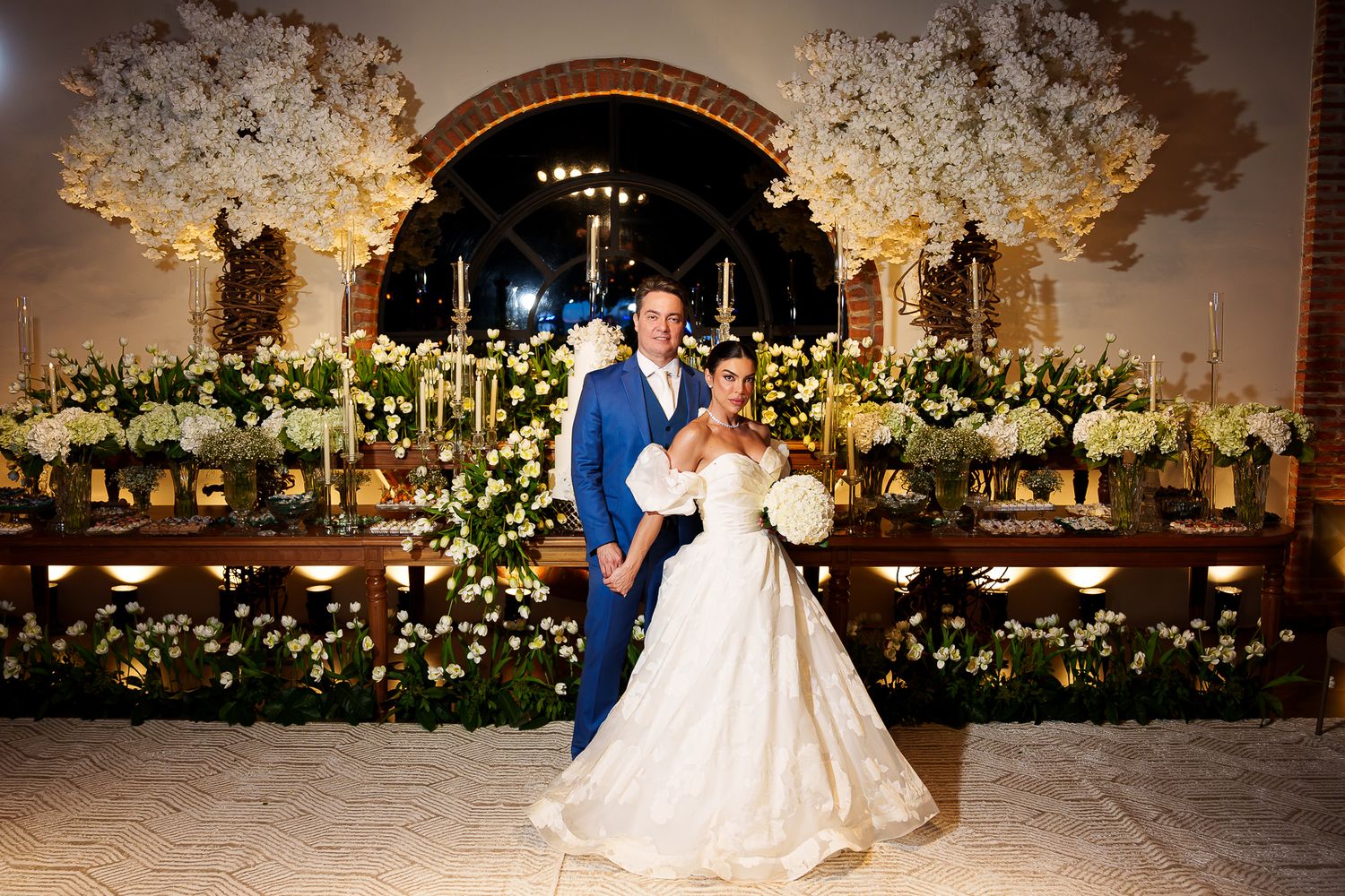 Bride in white ball gown and groom in blue suit pose before floral arch with white blooms at elegant wedding reception.