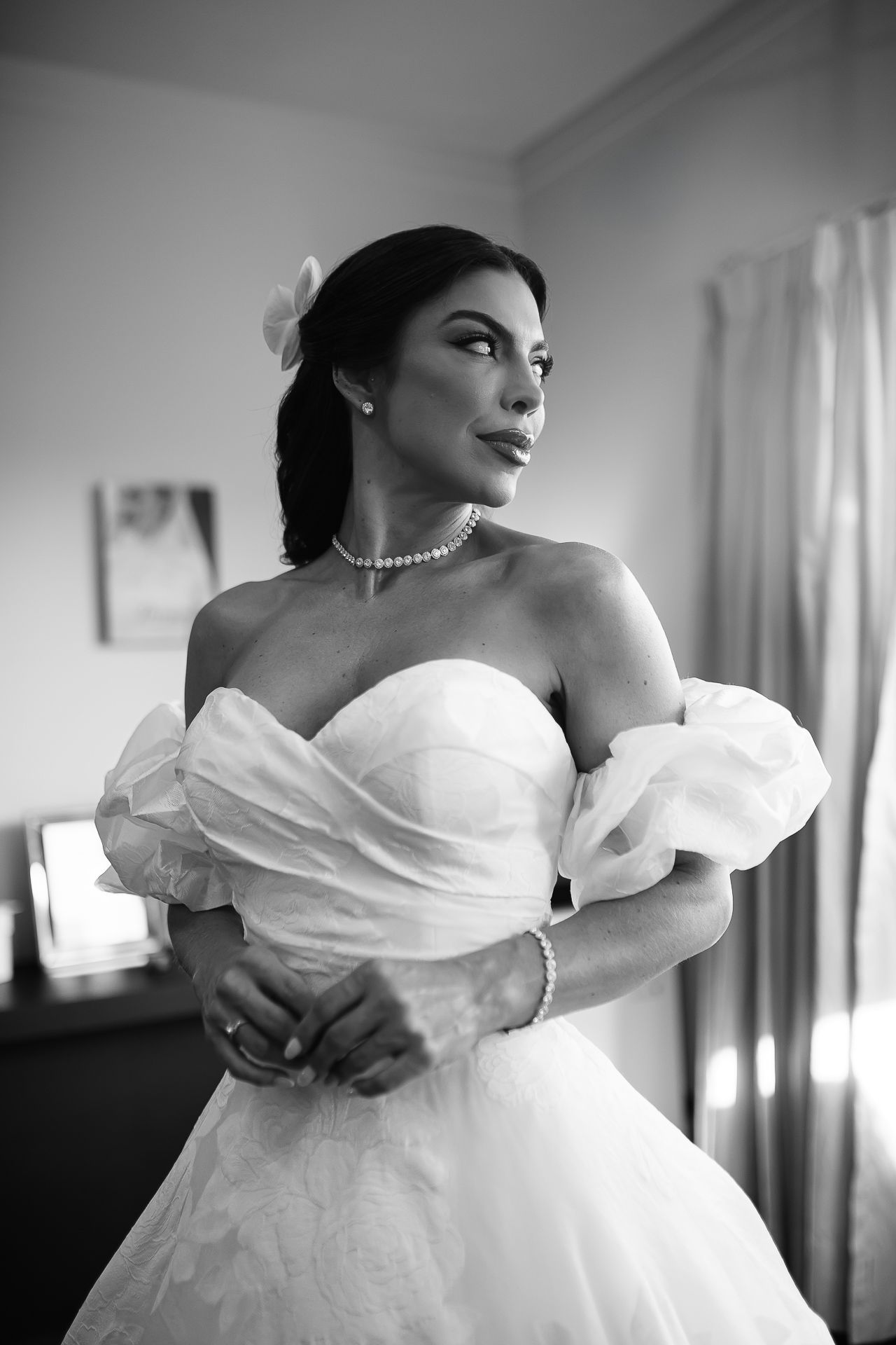 Black and white photo of a bride in an off-shoulder ball gown with pearl jewelry, posing elegantly indoors.