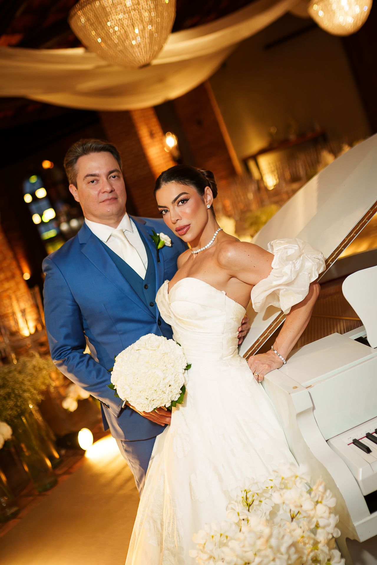 Elegant bride in white gown with bouquet and groom in blue suit pose by white piano at romantic wedding reception.