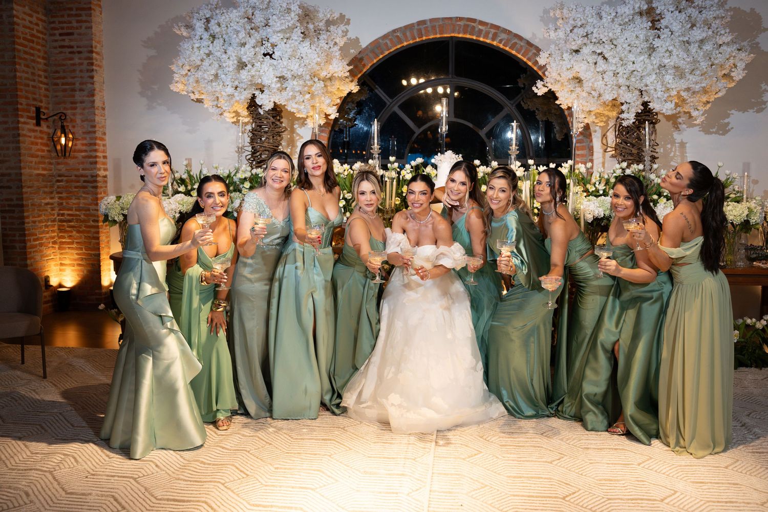 Bride in white gown poses with bridesmaids in green satin dresses at elegant floral wedding reception.