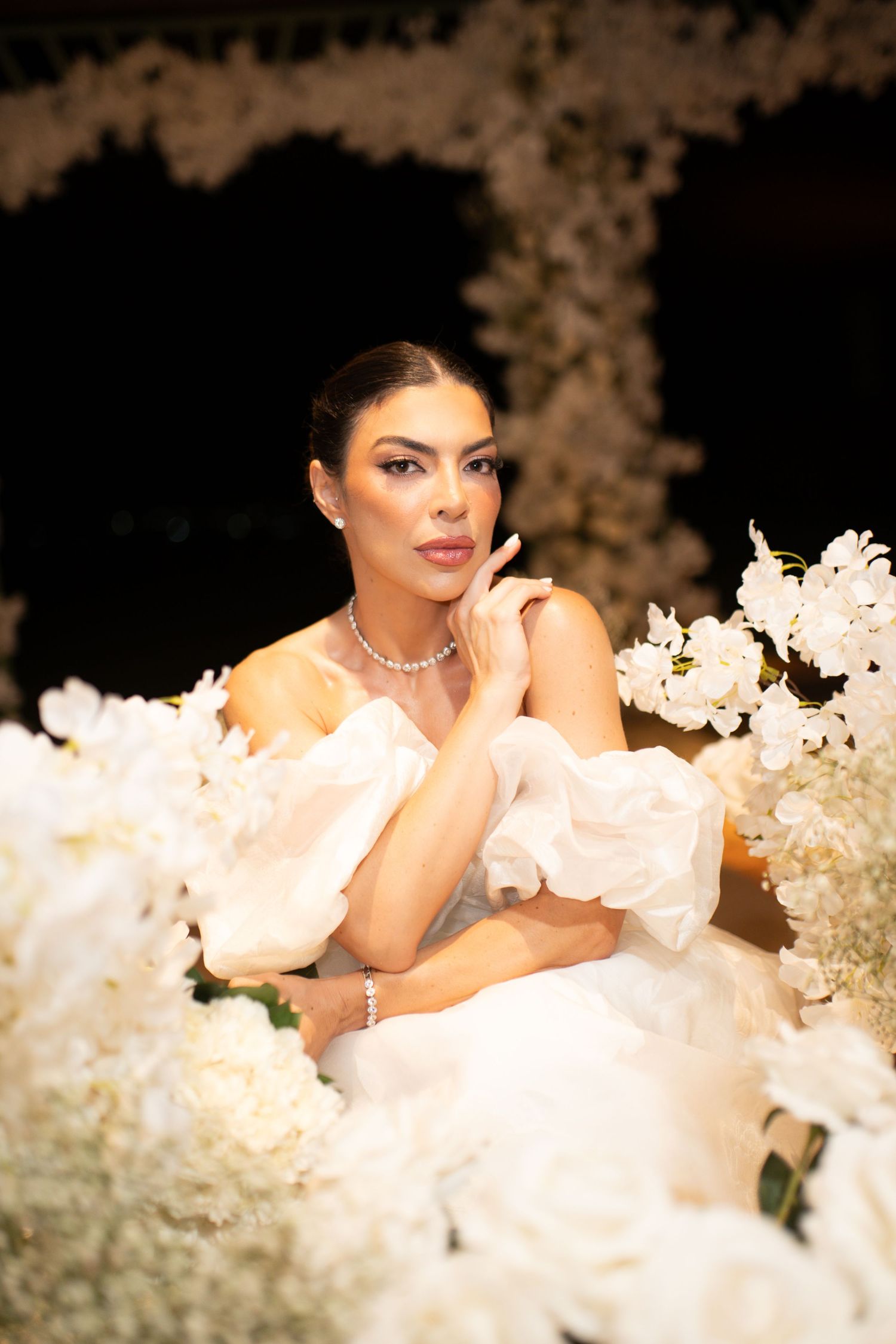 Elegant bride in white off-shoulder gown surrounded by white flowers, wearing diamond jewelry, posing gracefully.