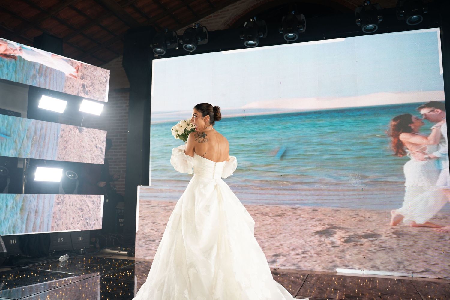 Bride in white off-shoulder ball gown holds bouquet before large beach screen display at elegant wedding venue.