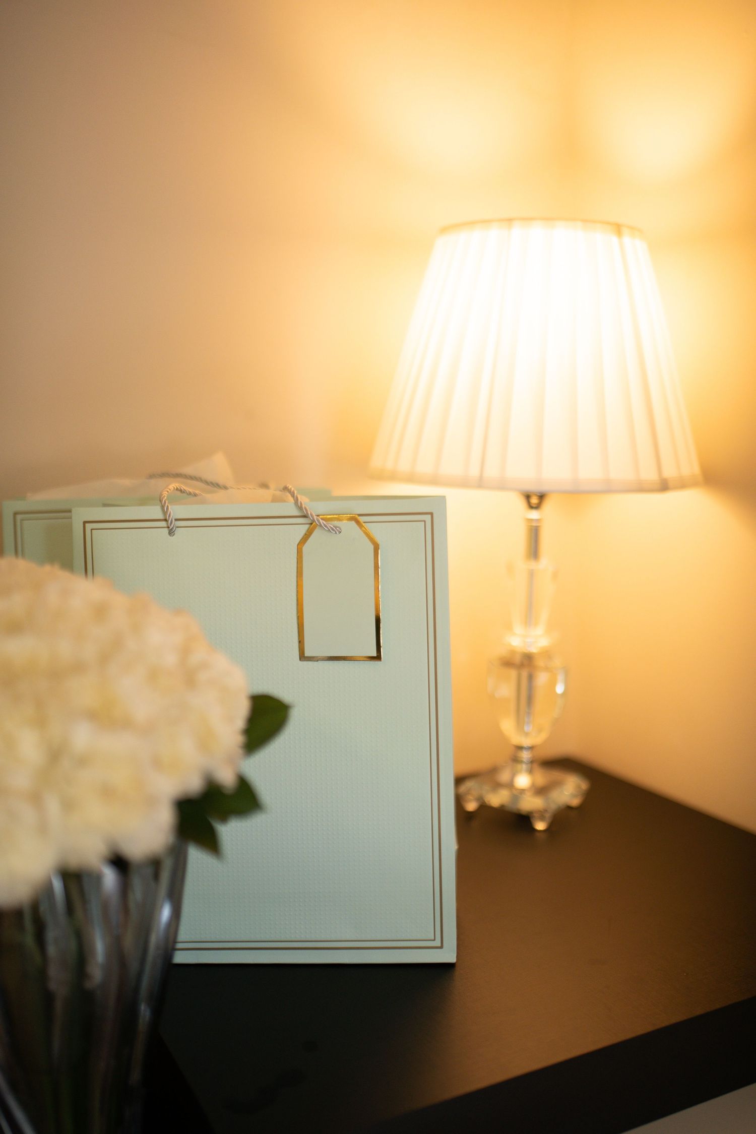 Elegant bedside table with glowing crystal lamp, white hydrangeas, and a gift bag with gold tag.