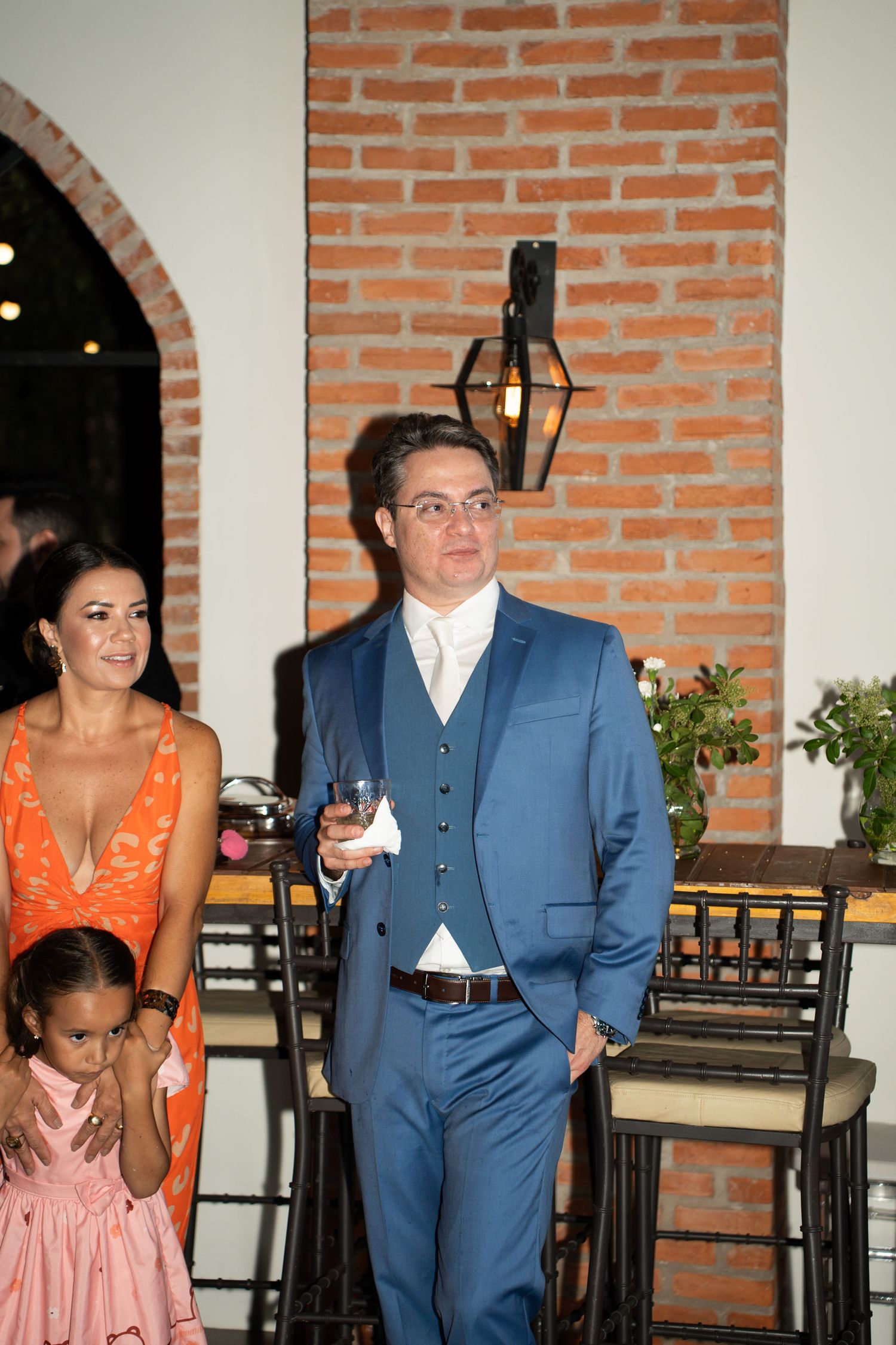 Man in blue suit holding drink at elegant wedding reception with woman in orange dress and child nearby.