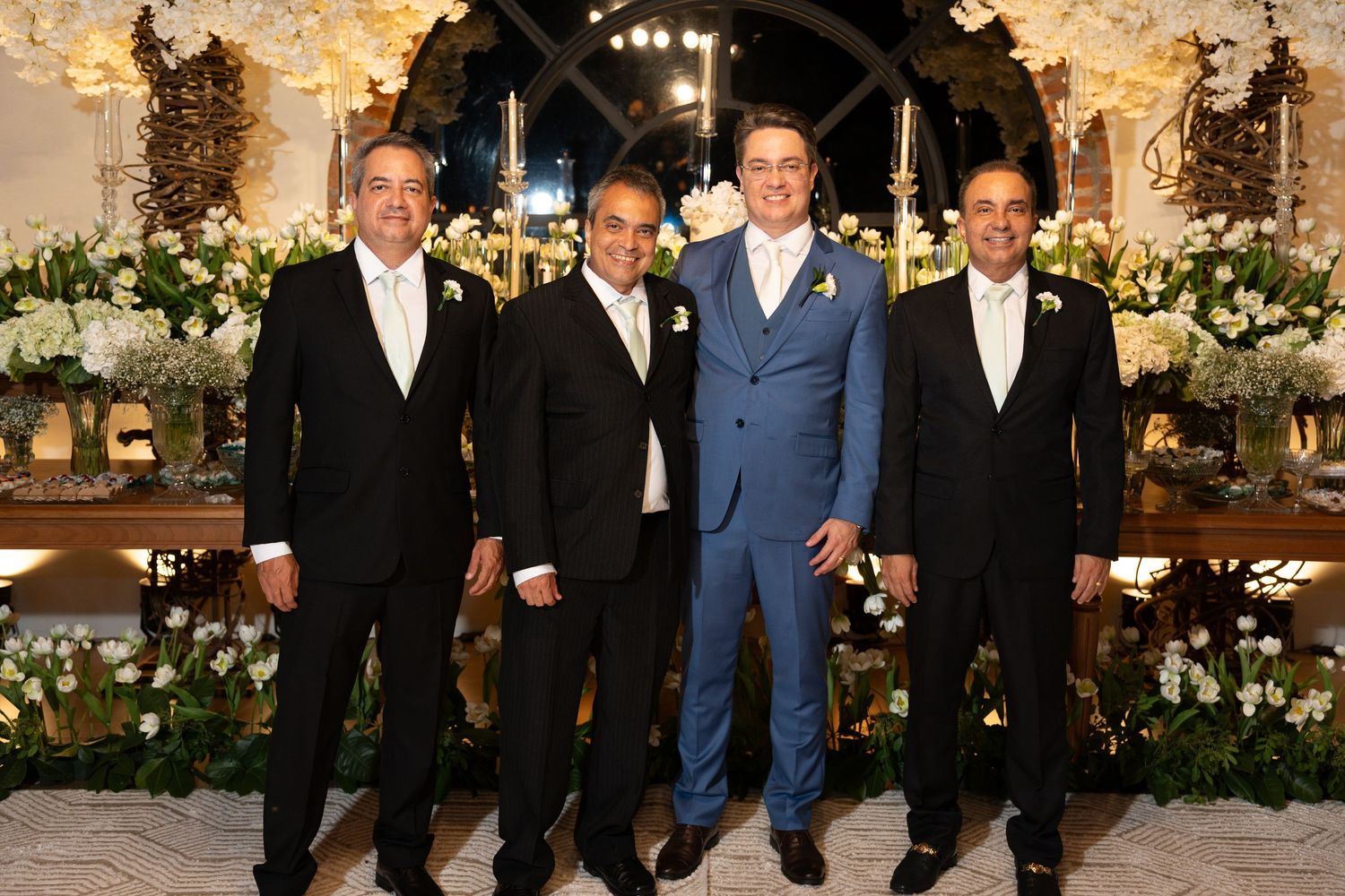 Four men in formal suits posing together at an elegantly decorated wedding venue with white floral arrangements.