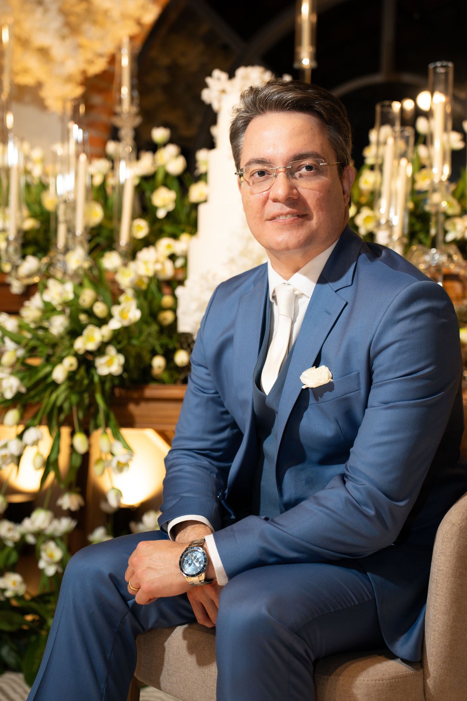 Elegant man in blue suit with white pocket square seated before floral wedding backdrop with candles.
