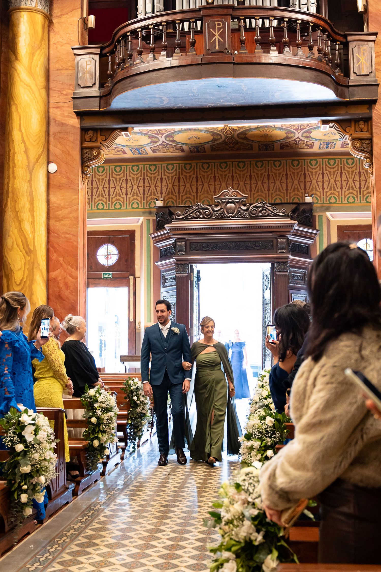 Couple walks down the aisle of an ornate venue lined with white floral arrangements during their wedding ceremony.