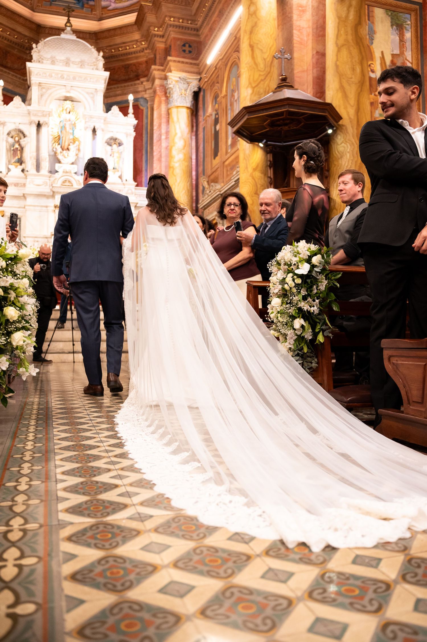 Bride in cathedral-length veil walks down church aisle with groom during elegant wedding ceremony.