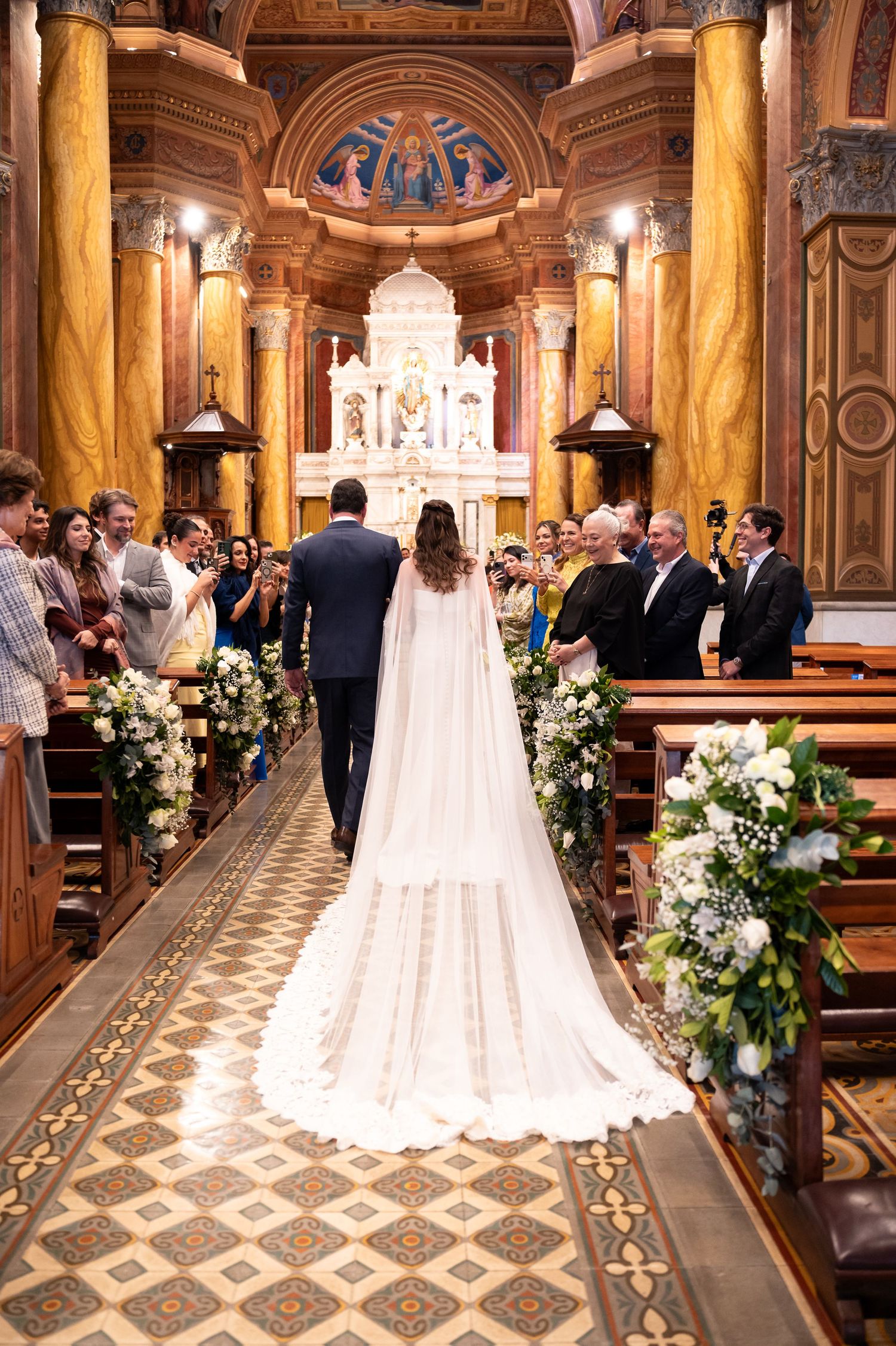 Bride in white gown with long cape walks down church aisle with groom toward ornate altar adorned with flowers.