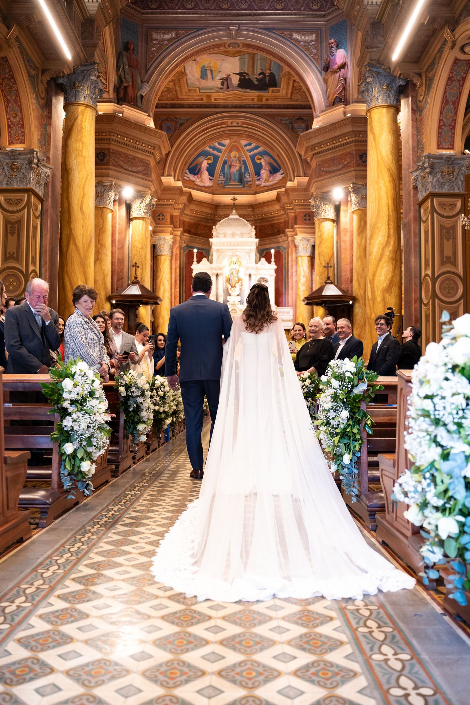 Bride and groom walk down ornate church aisle lined with white floral arrangements during elegant wedding ceremony.