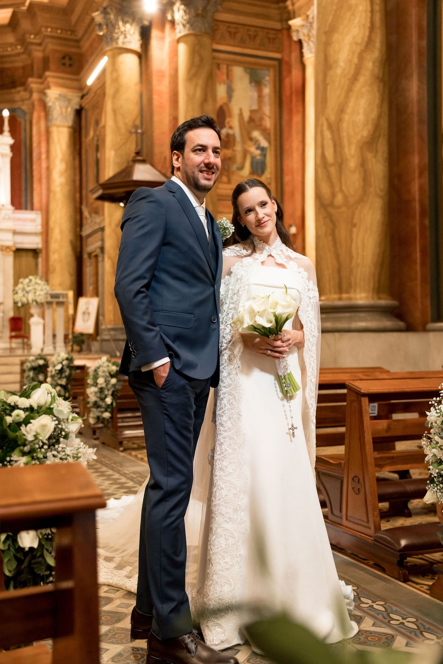 Bride and groom posing together in an ornate church, bride holding white bouquet wearing lace gown with cape.