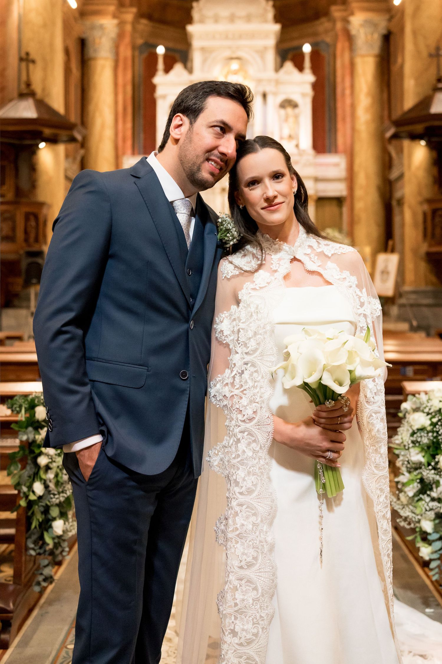 Elegant bride and groom posing inside a church, bride wearing lace cape gown holding calla lily bouquet, groom in navy suit.