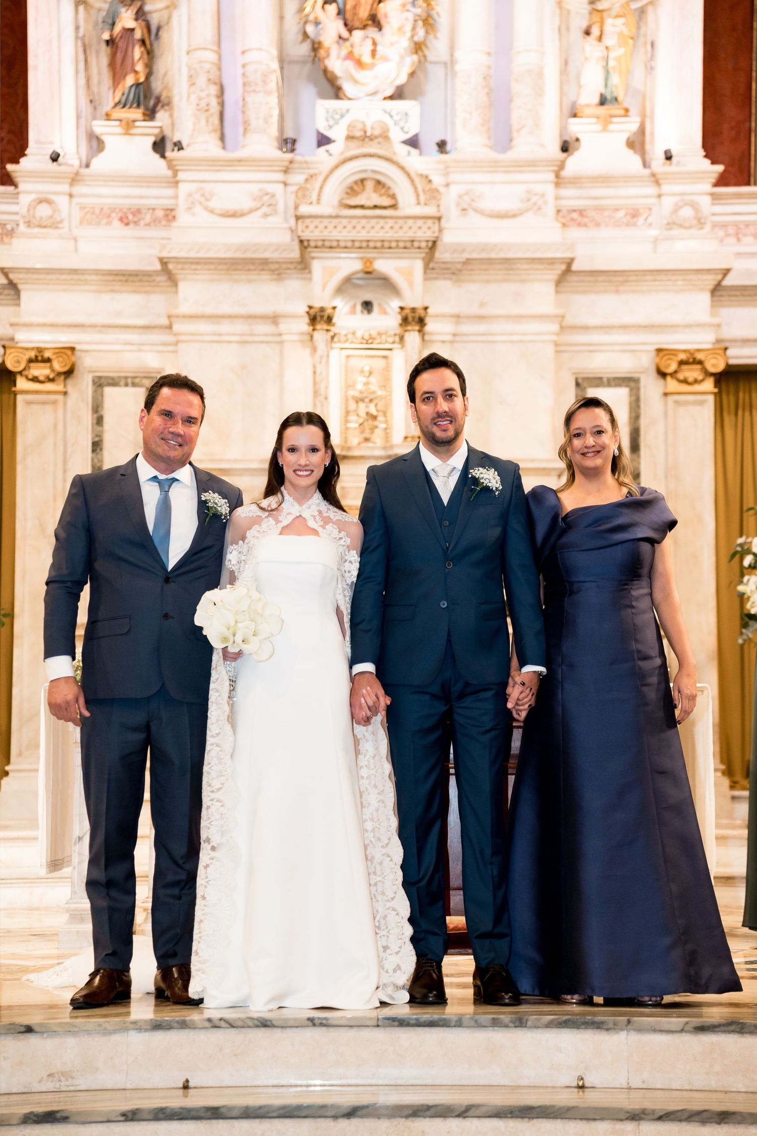 Wedding party of four posing at ornate church altar, bride in white lace gown with bouquet, groom and guests in navy attire.